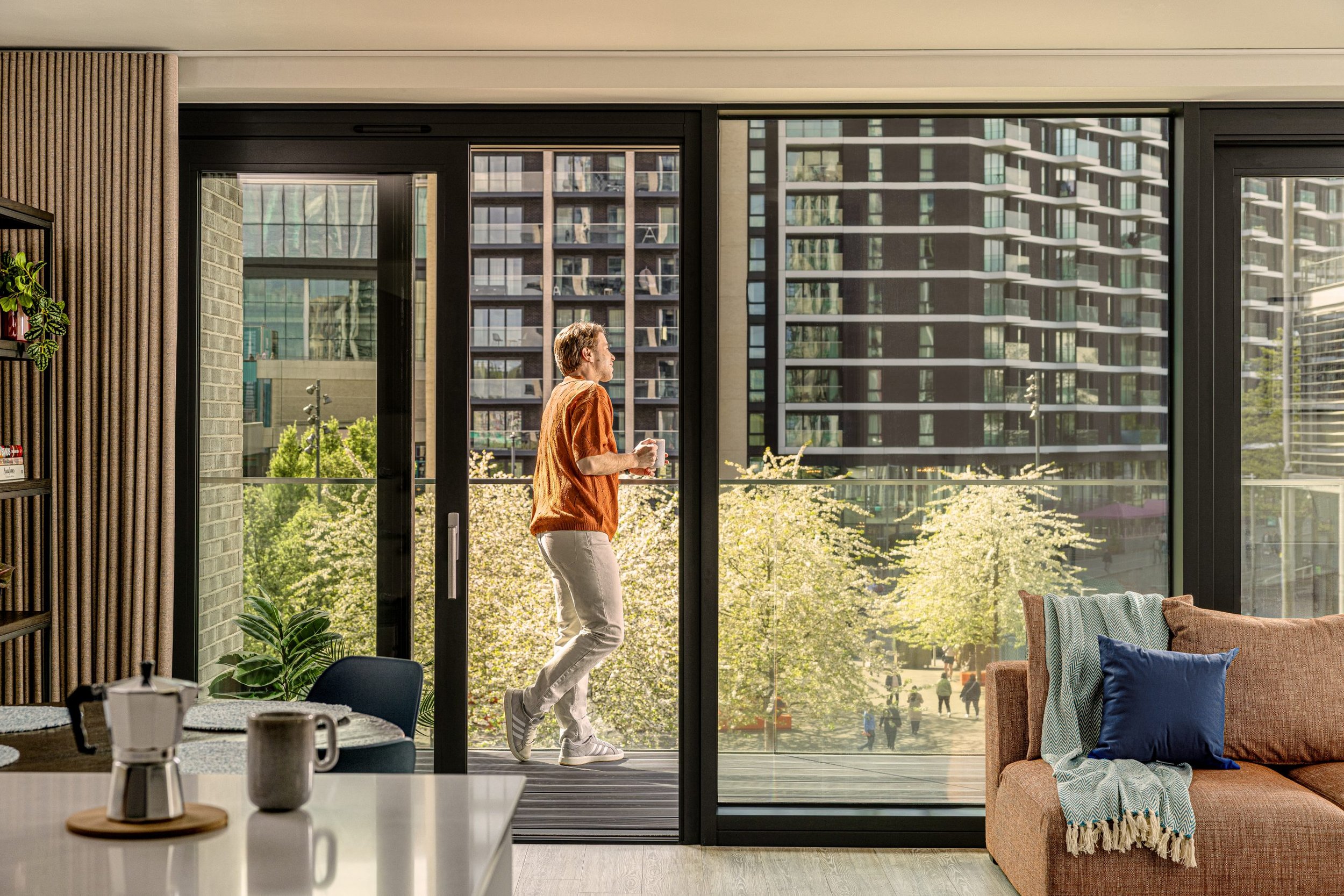 A person in an orange jacket and white pants is walking and holding a coffee mug near a large glass sliding door in an urban apartment, with high-rise buildings outside.