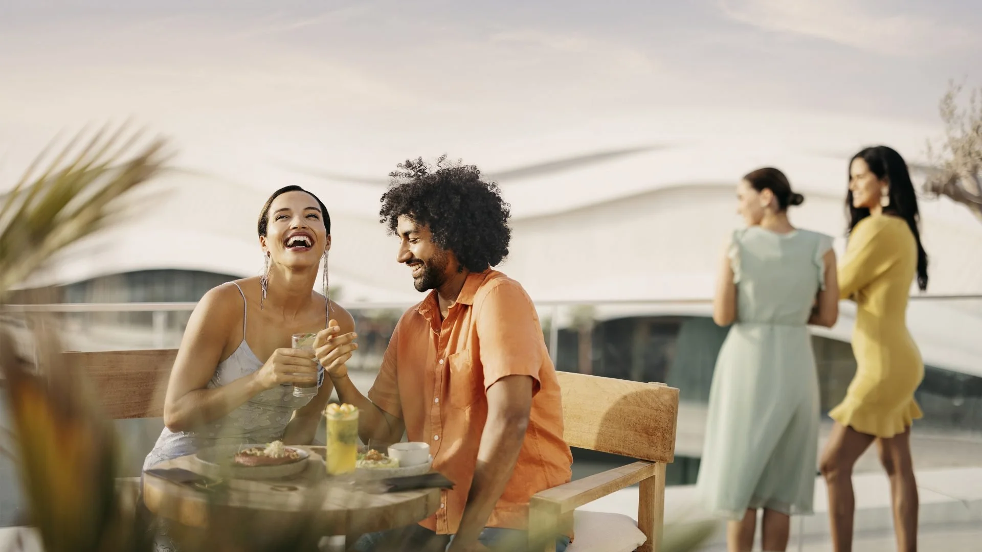 Two people laughing and chatting at an outdoor restaurant table with food and drinks, while two women in dresses stand and talk in the background under a canopy.