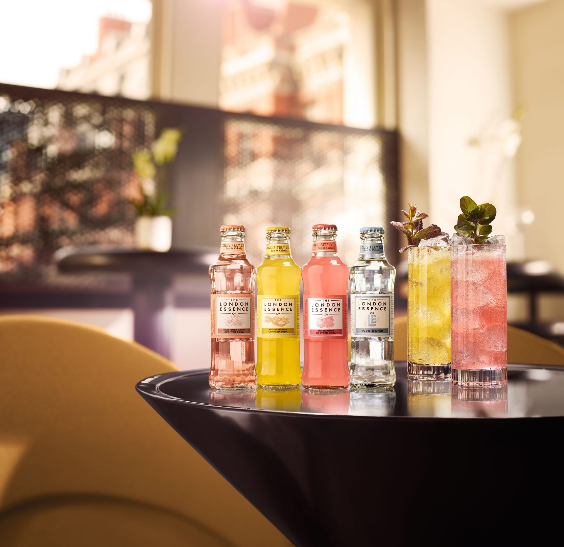 Four bottles of craft soda in pink, yellow, red, and clear flavors are arranged on a black tray, with a glass filled with yellow and pink soda and garnished with mint and herbs, in front of a window with a blurred urban background.