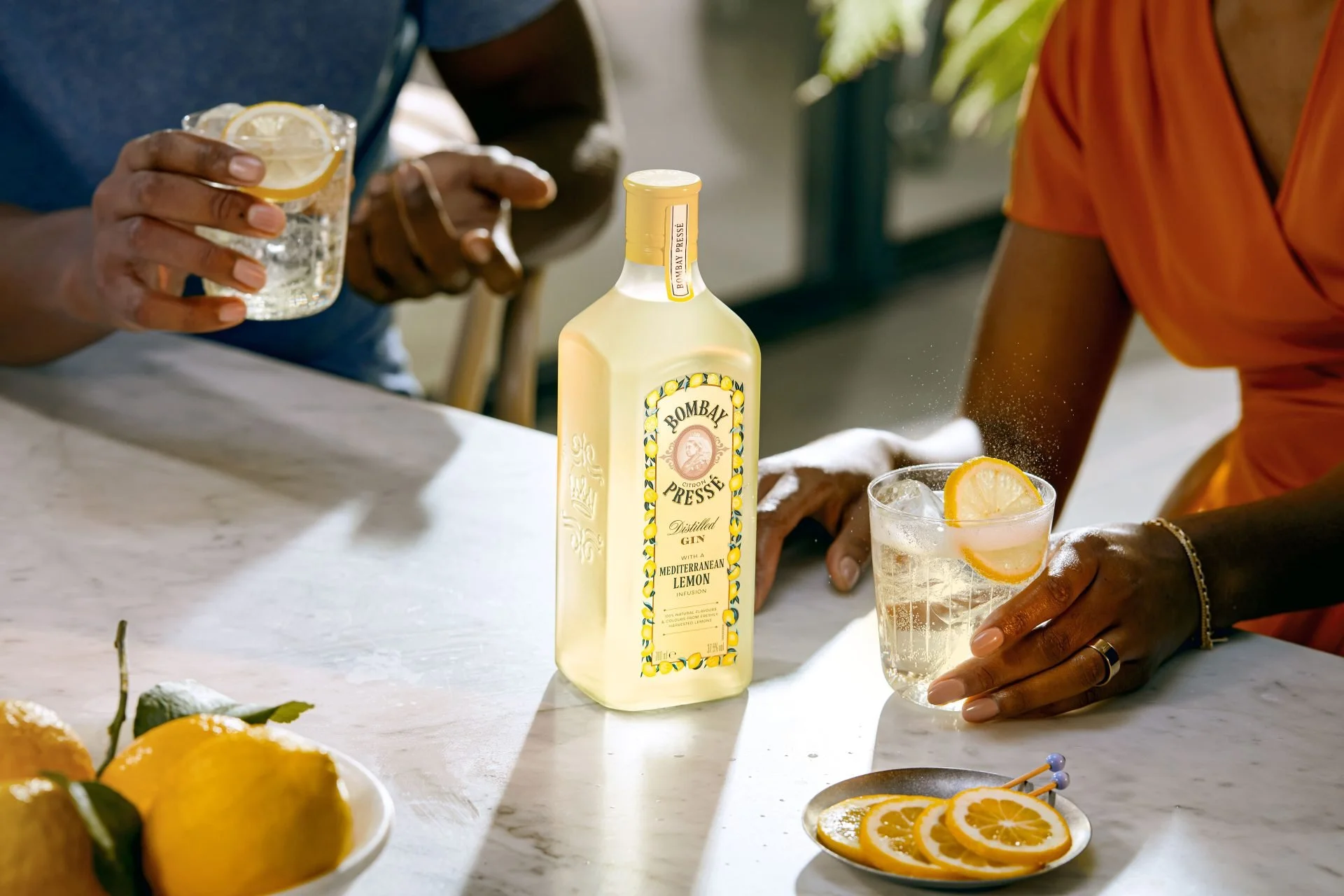 A close-up of a bottle of Bombay Presse Mediterranean Lemon Gin on a white marble table, with two people serving lemon gin cocktails in glasses garnished with lemon slices. One person is holding a glass with lemon slices, and another person is reachi