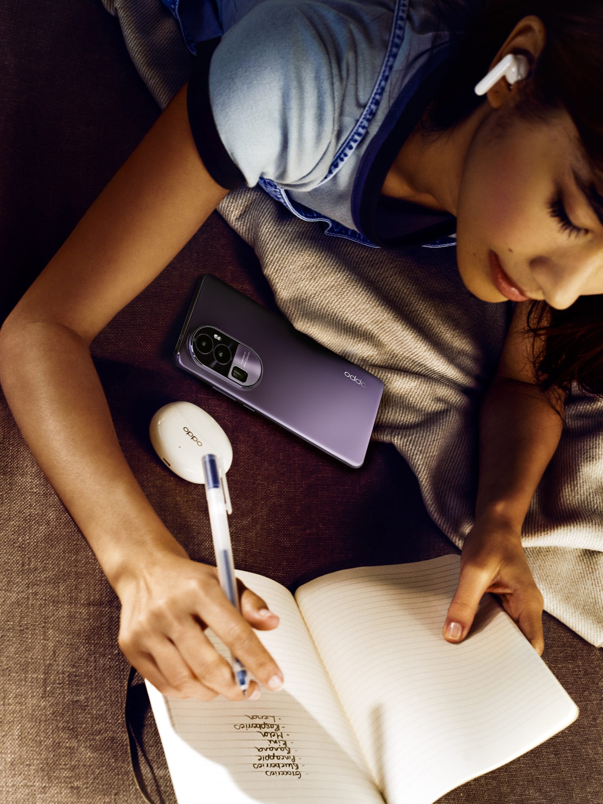 A woman is writing in a notebook with a blue pen. She has a smartphone, wireless earbuds, and a white wireless earbud case on her lap. She is sitting on a couch, and her left hand is pointing to a list of words in the notebook.