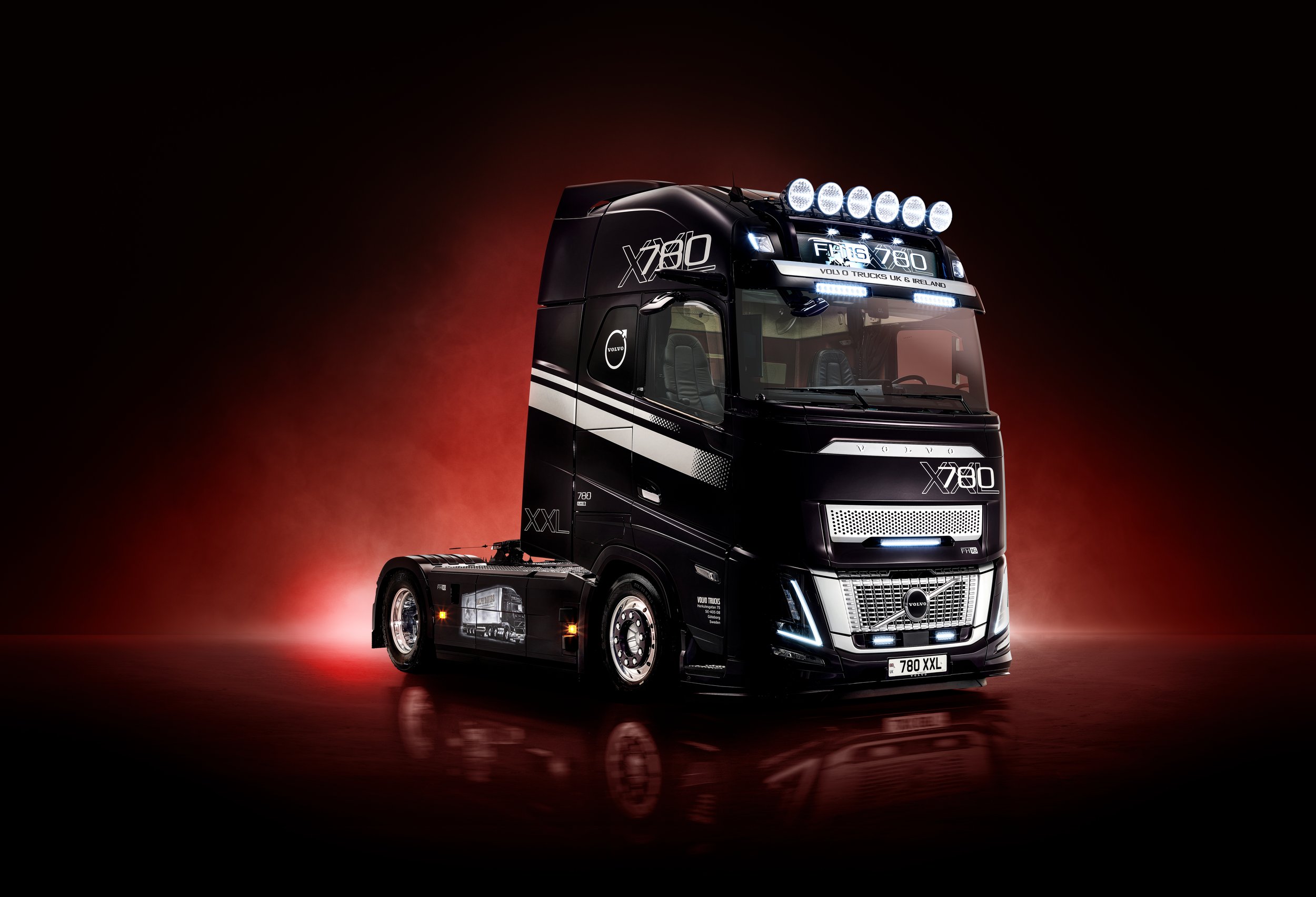 A black Volvo truck with bright lights on the roof and front grille, set against a dark background with a red glow, and a reflection on the shiny floor.