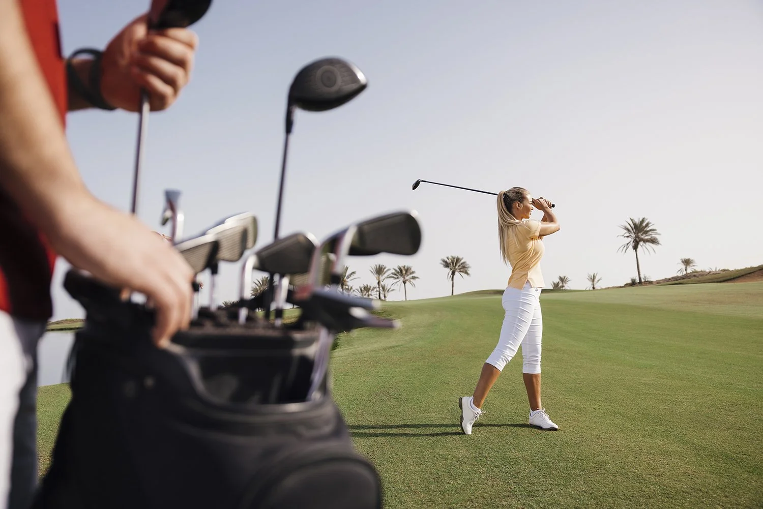 A woman swinging a golf club on a golf course with palm trees in the background, while a person with a golf bag watches.