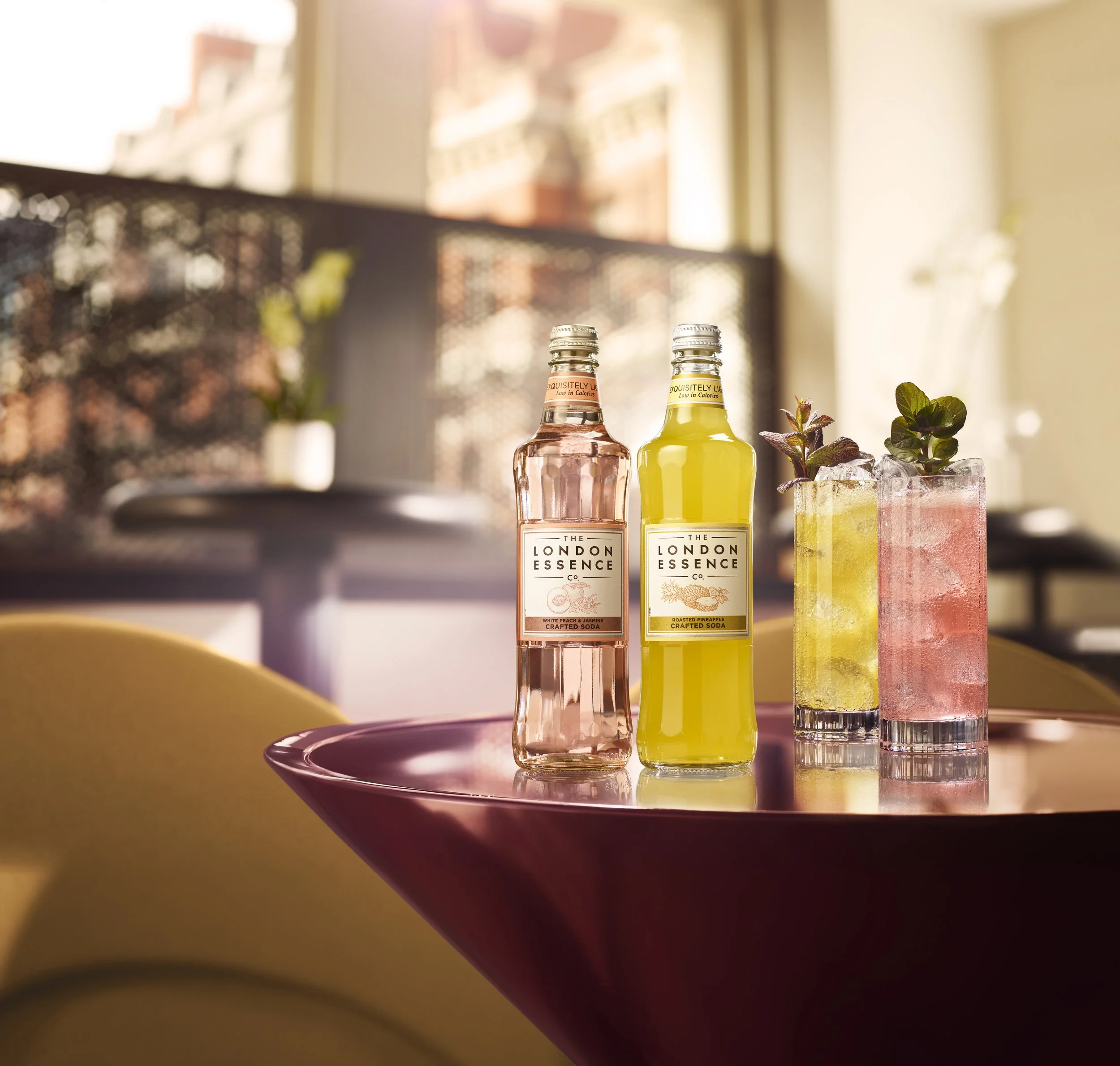 Two glass bottles on a pink tray, with greenery in a clear glass beside them, on a purple table near a window with a blurred background of buildings.