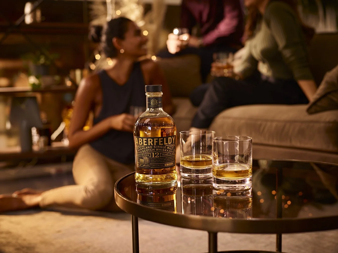 A bottle of Aberfeldy Highland Single Malt Scotch Whisky with two glasses of whiskey on a round black table in a cozy, dimly lit lounge where people are socializing in the background.