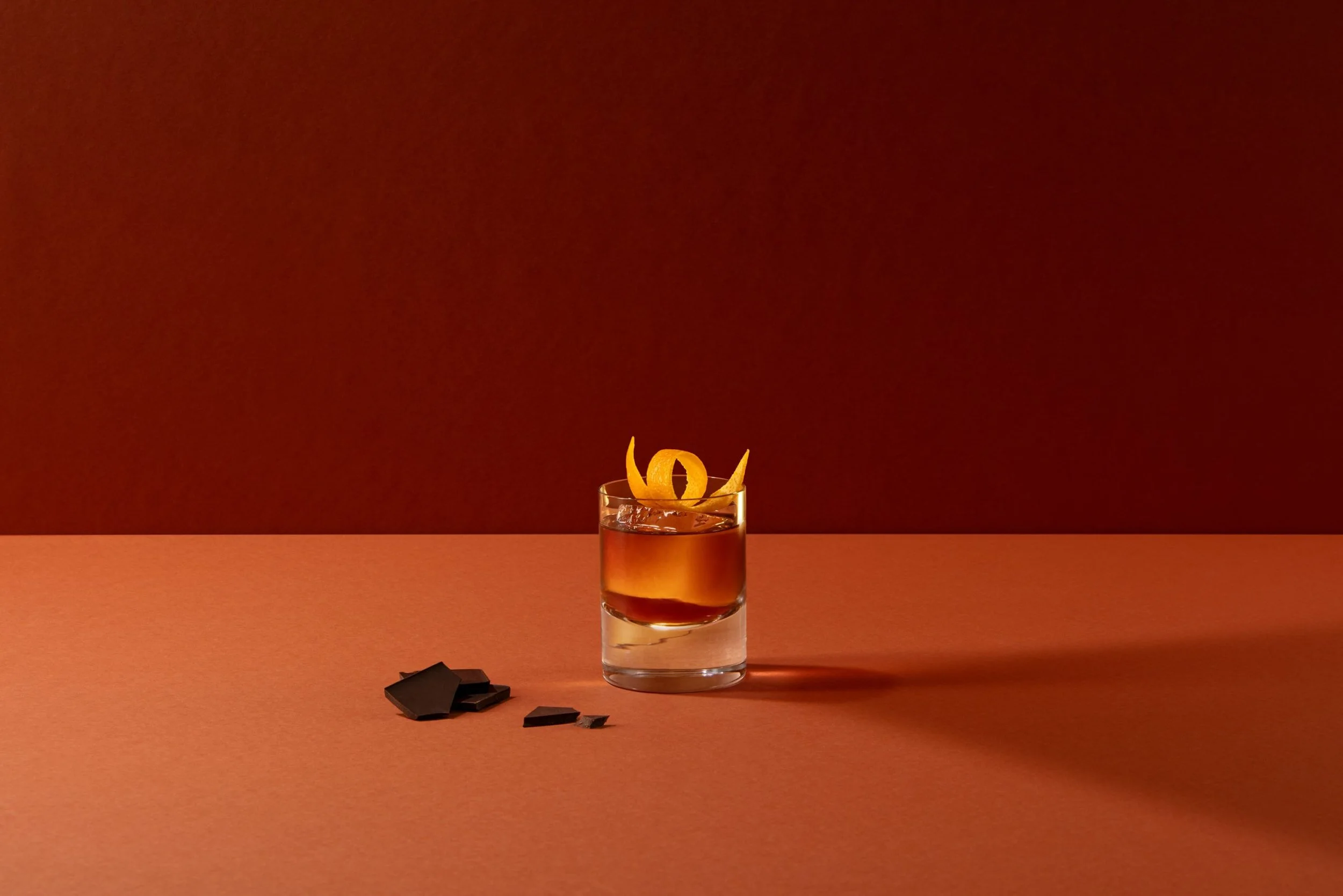 A short glass of dark red beverage garnished with curled orange peels on a terracotta-colored surface against a matching warm terracotta background.