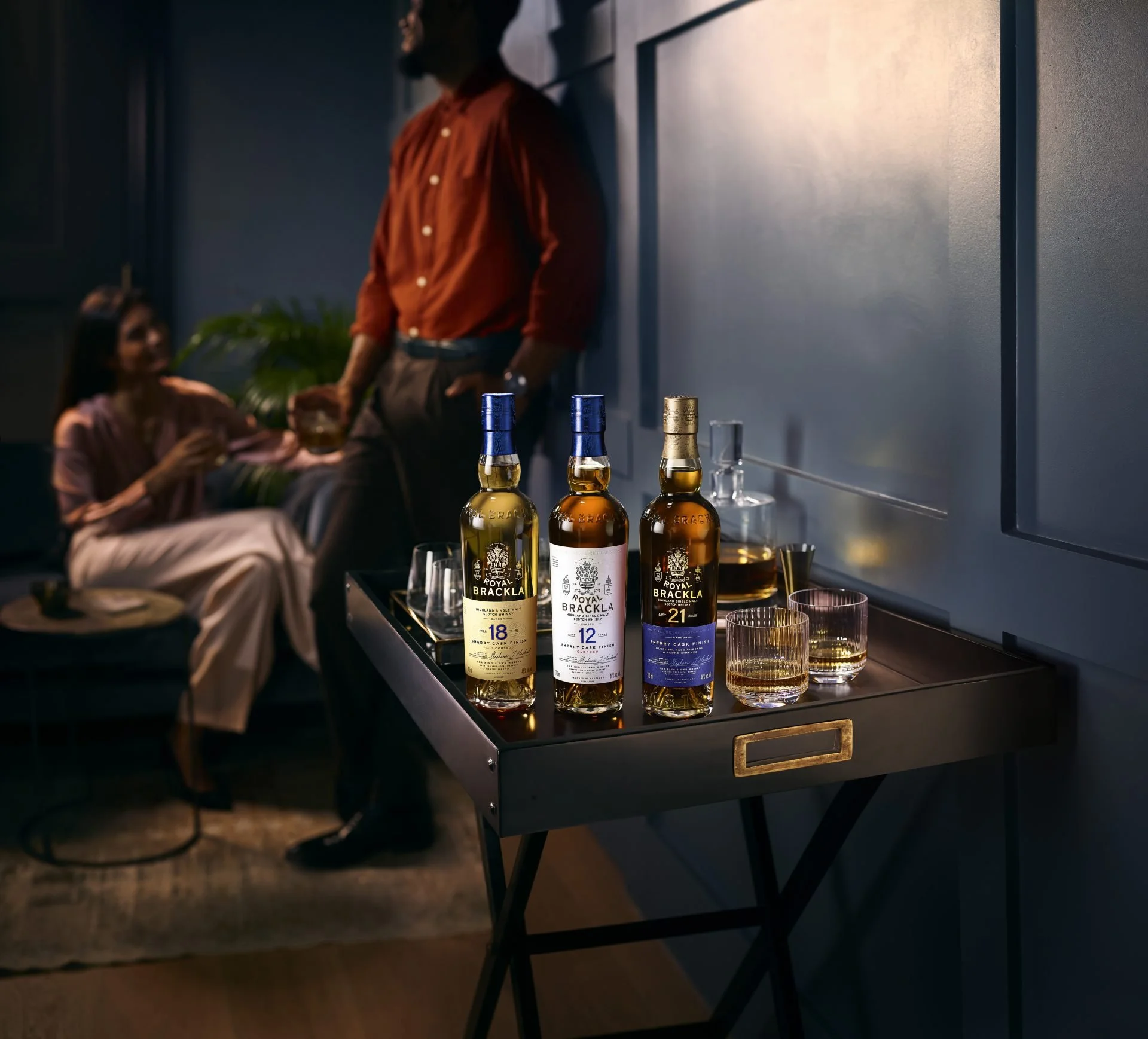 Three bottles of Royal Brackla whisky on a black table with glasses filled with whisky and ice in front of a blue wall. Two people are in the background, one sitting and using a smartphone, the other standing and talking, both wearing casual attire.