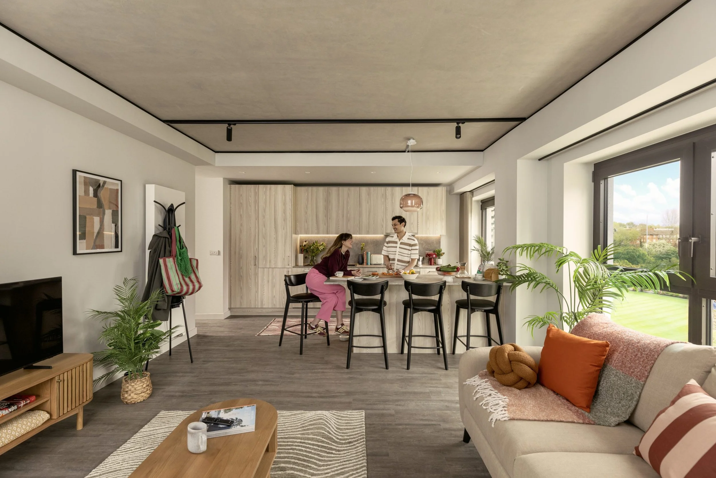 A modern living room with large windows, a beige sofa with orange and striped throw pillows, and a kitchen area in the background with two people cooking beside a kitchen island. There are plants, a wooden coffee table, and wall art.