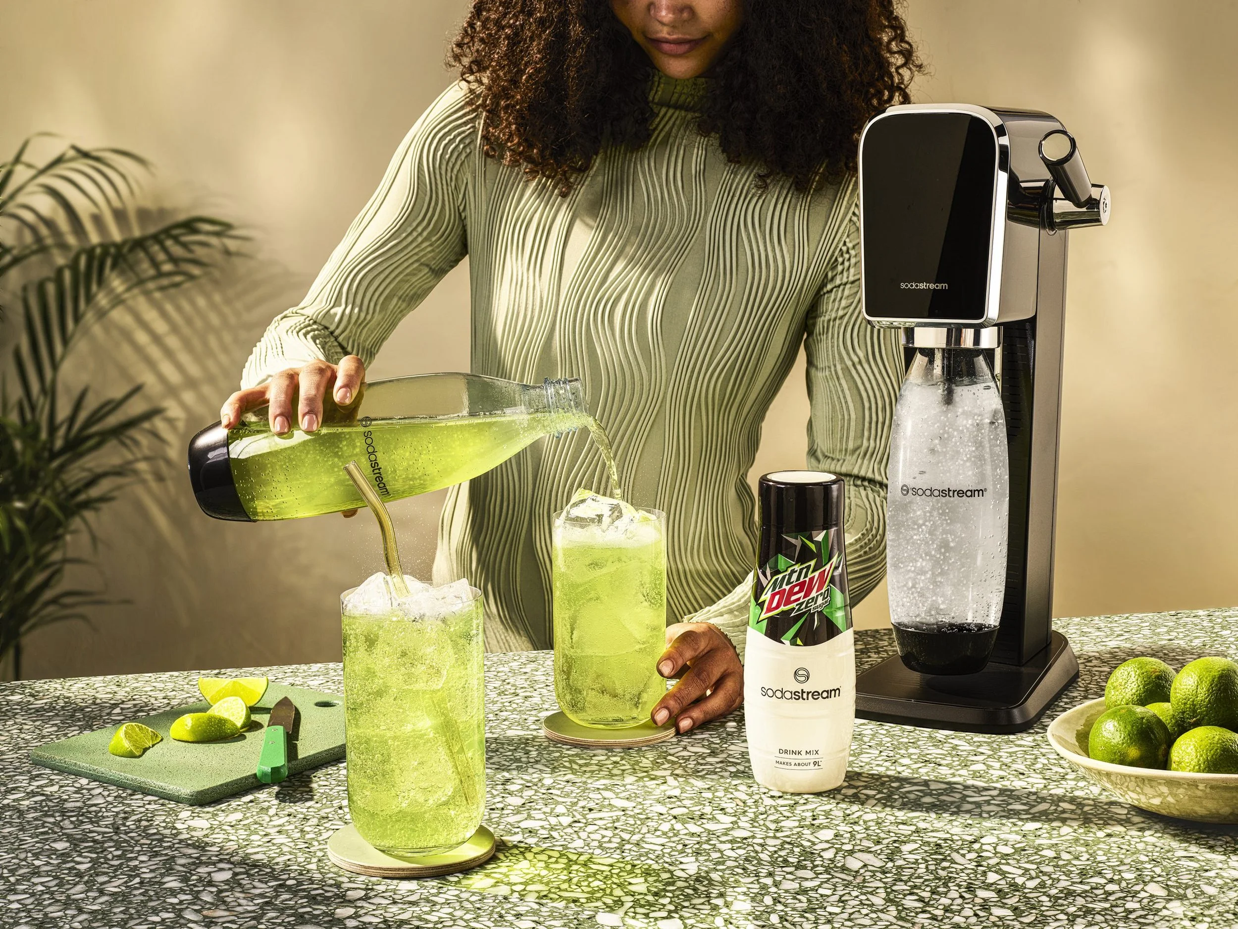 A woman with curly hair in a green striped sweater is pouring green lime-flavored drink from a SodaStream machine into a glass over ice. On the black and white speckled countertop, there are lime wedges, a sliced lime, a bottle of Mountain Dew Zero, 