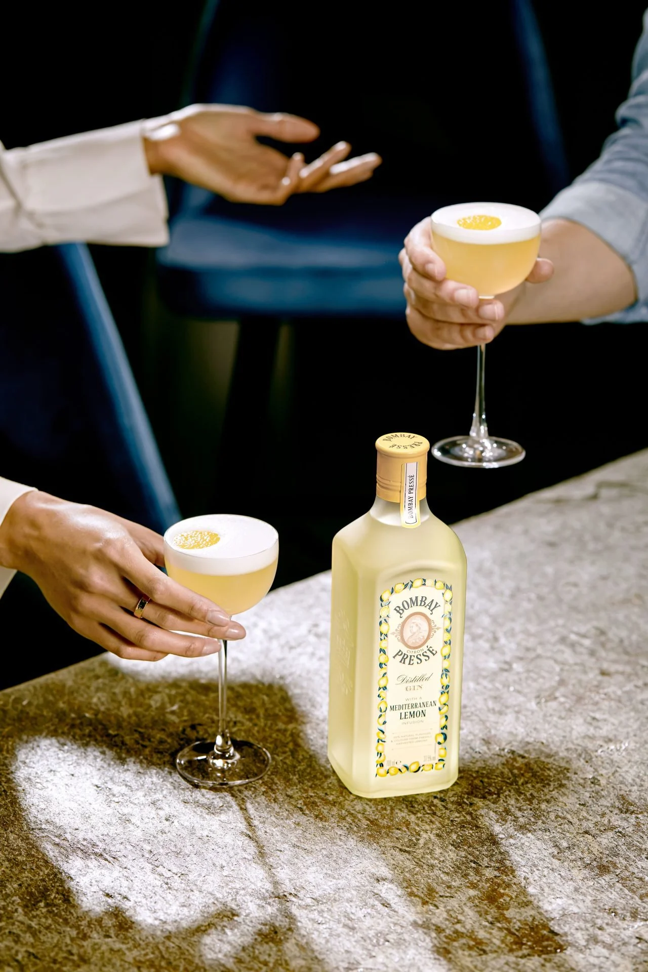 Two people are holding cocktail glasses filled with a yellow drink topped with frothy foam, near a bottle of Bombay Presse Mediterranean Lemon gin on a textured surface.