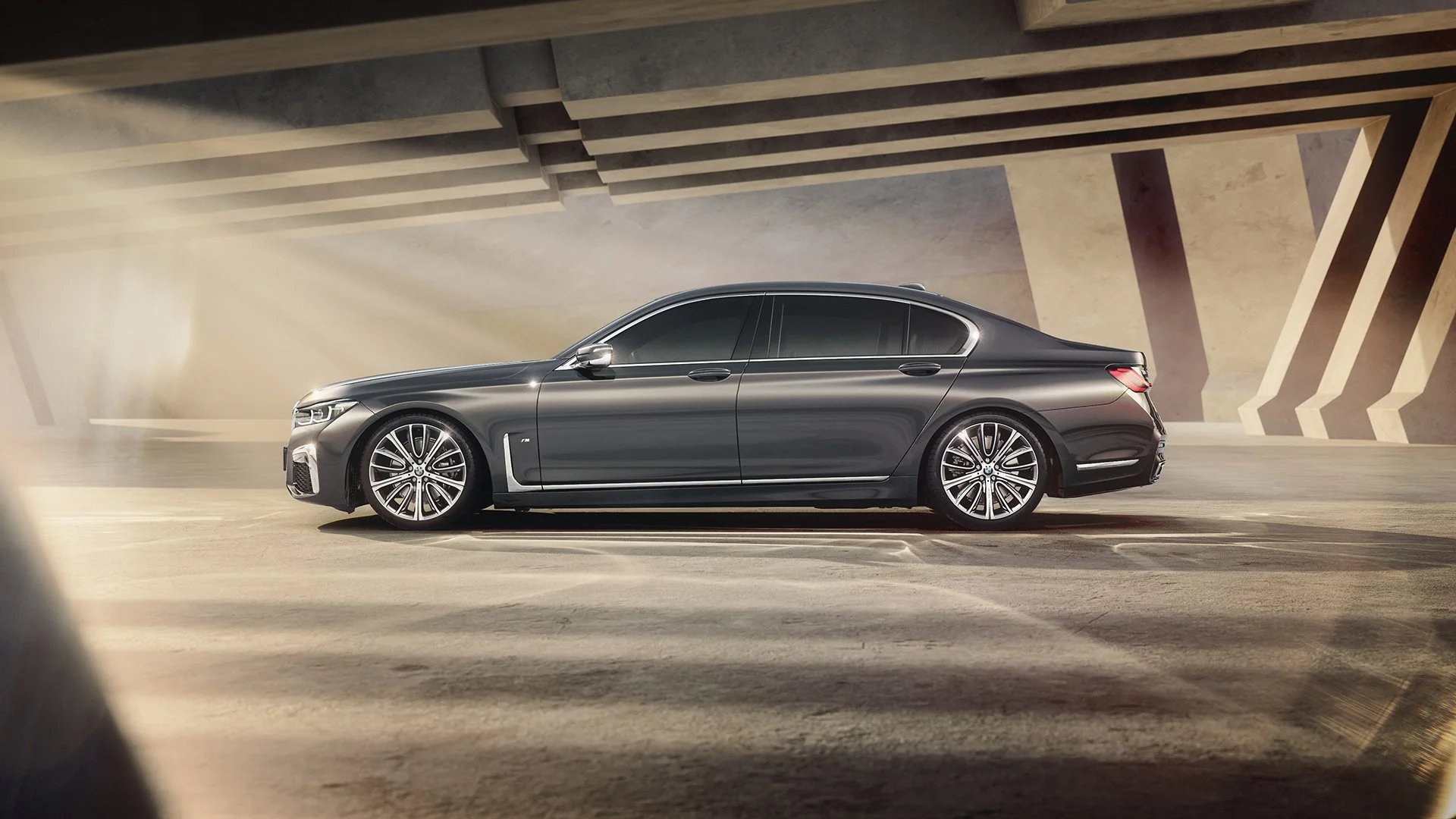 A sleek, dark gray luxury sedan parked under an architecturally modern structure with geometric design. Sunlight shines on the car, highlighting its smooth curves and stylish alloy wheels.