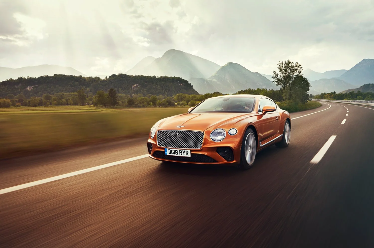 Orange luxury sports car driving on a curved mountain road surrounded by green fields and mountains under a partly cloudy sky.