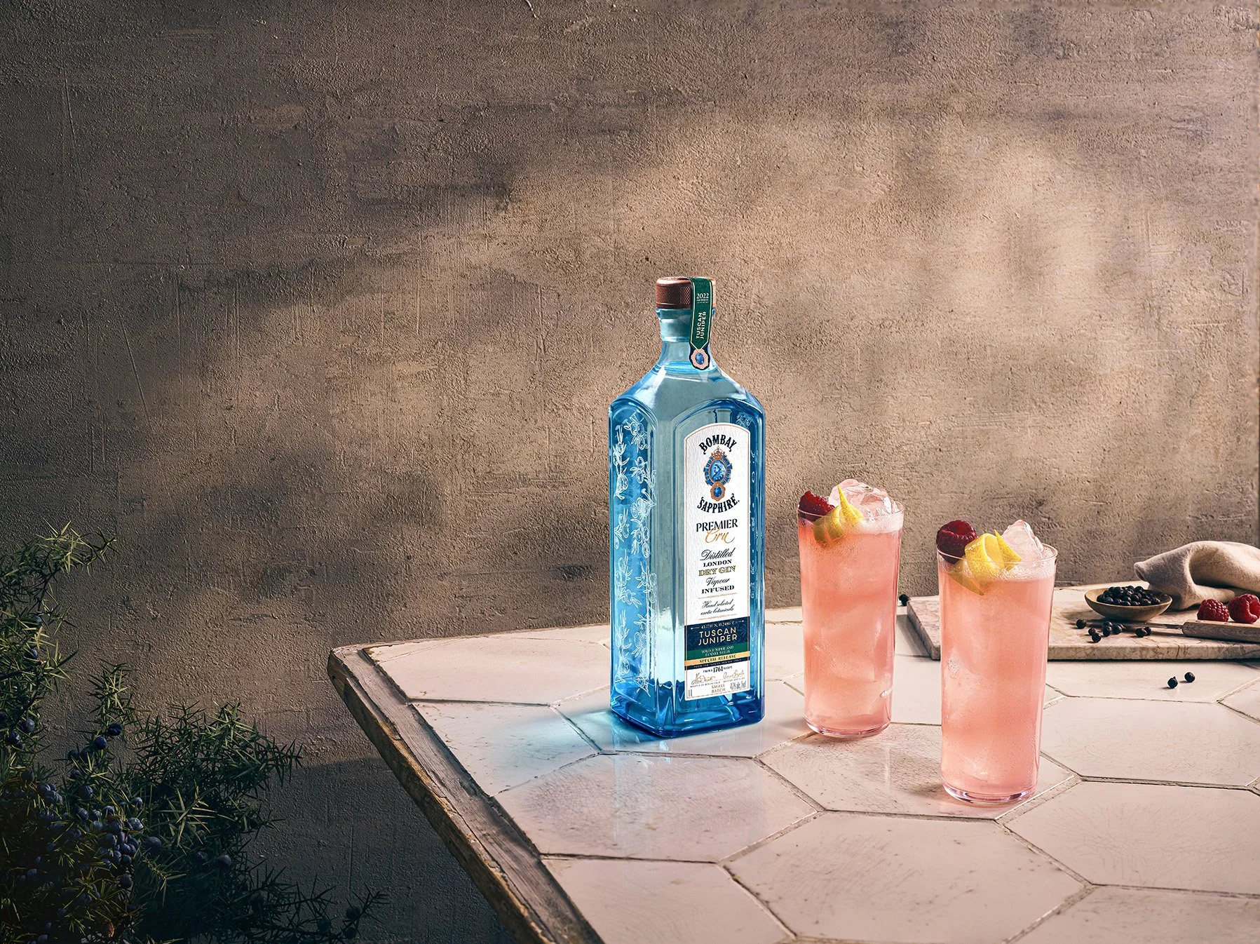 A bottle of Bombay Sapphire gin and two pink cocktails with lemon and raspberry garnish on a white tiled table, with a small bowl of blueberries and raspberries, and herbs in the background against a textured beige wall.