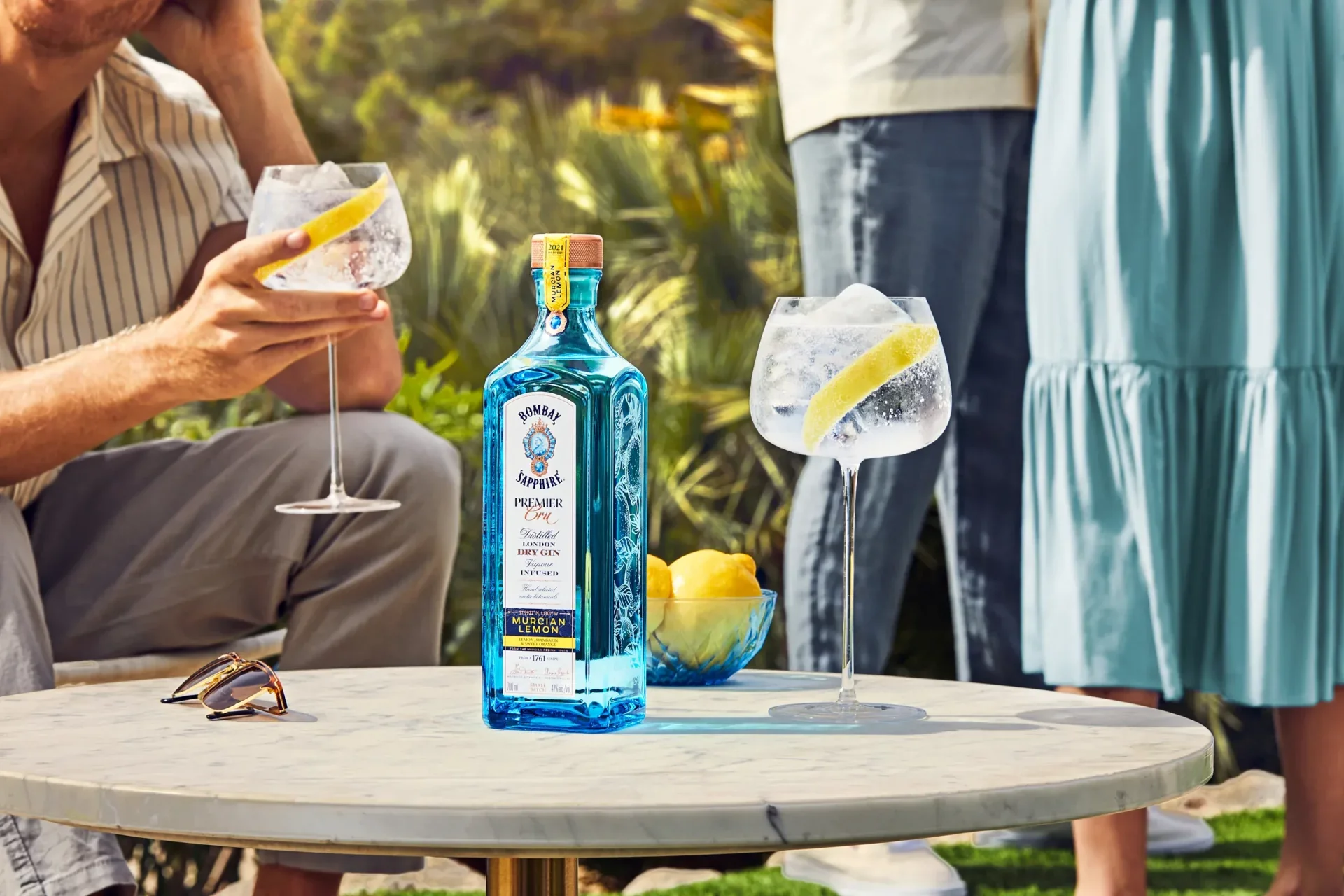 A bottle of Bombay Sapphire gin with a lemon wedge on the rim of a large glass filled with ice and a lemon wedge. Other glasses with ice and lemon wedges are on a table. Sunglasses and a bowl of lemons are also on the table. People are standing and s