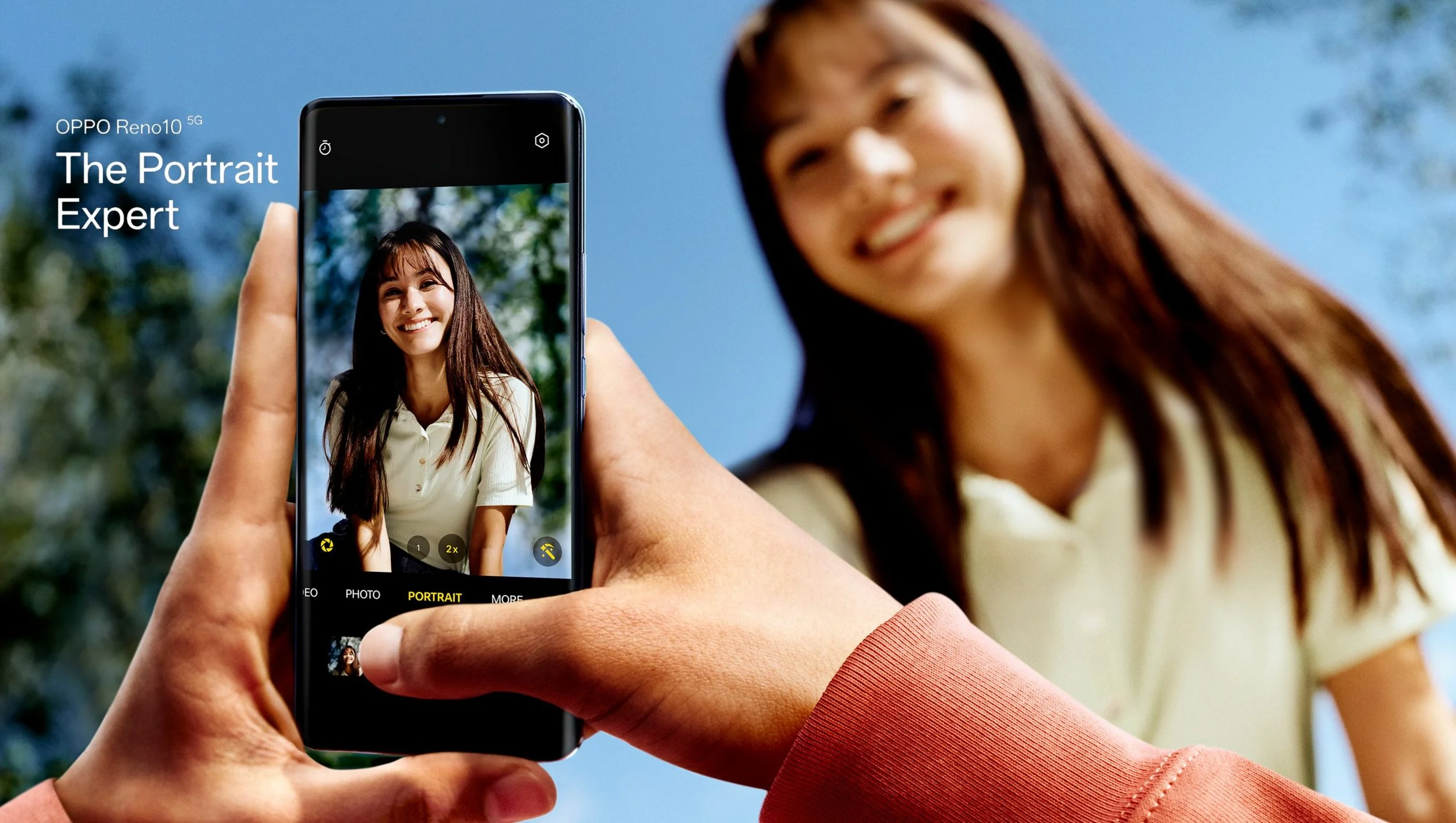 A person taking a photo of a smiling woman outdoors with a smartphone, with a blue sky and trees in the background.