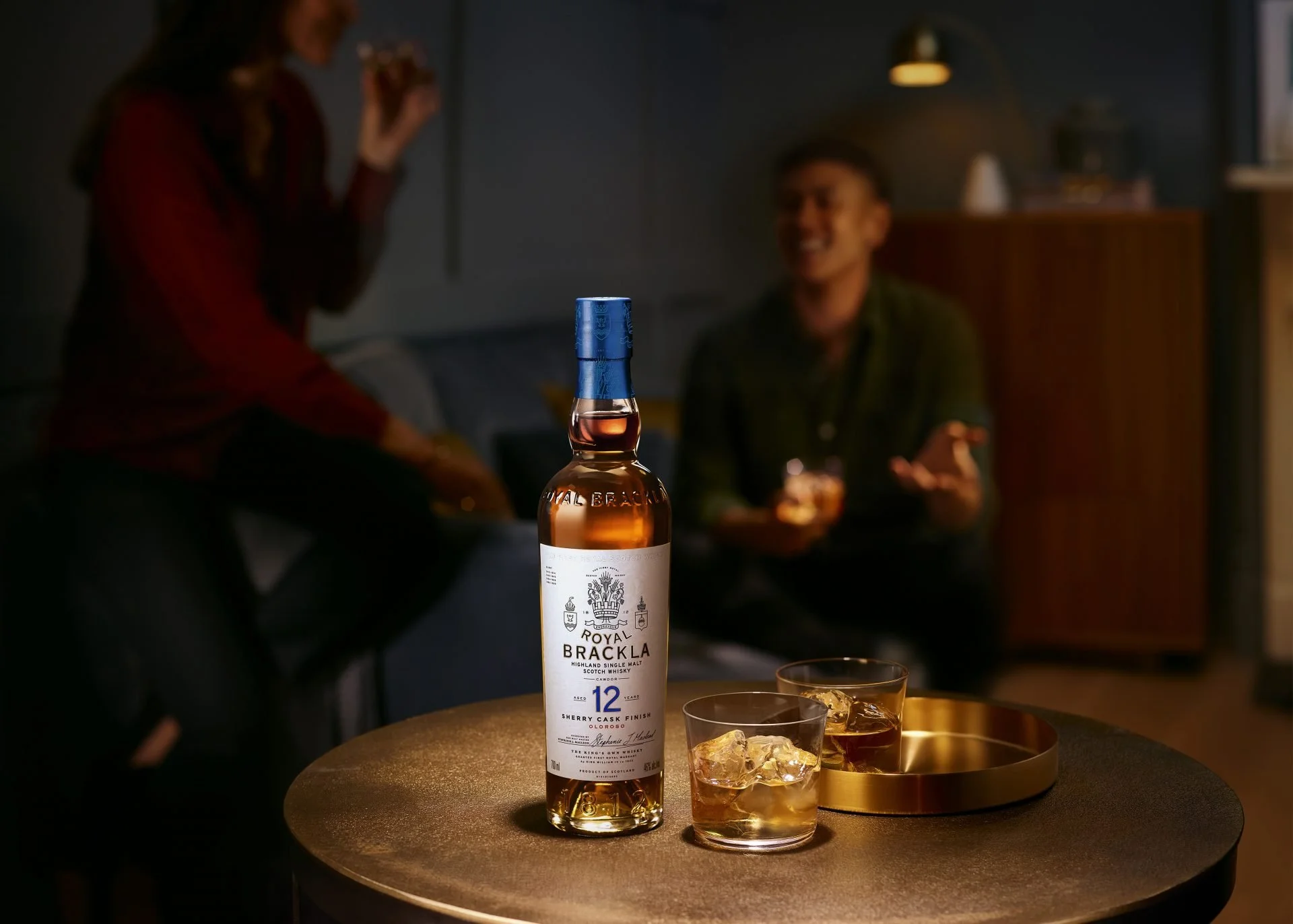 A bottle of Royal Brackla Highland Single Malt Scotch Whiskey with 12 years aged, labeled 'Sherry Cask Finish,' on a table with two glasses of whiskey and ice, in a dimly lit room with people socializing in the background.