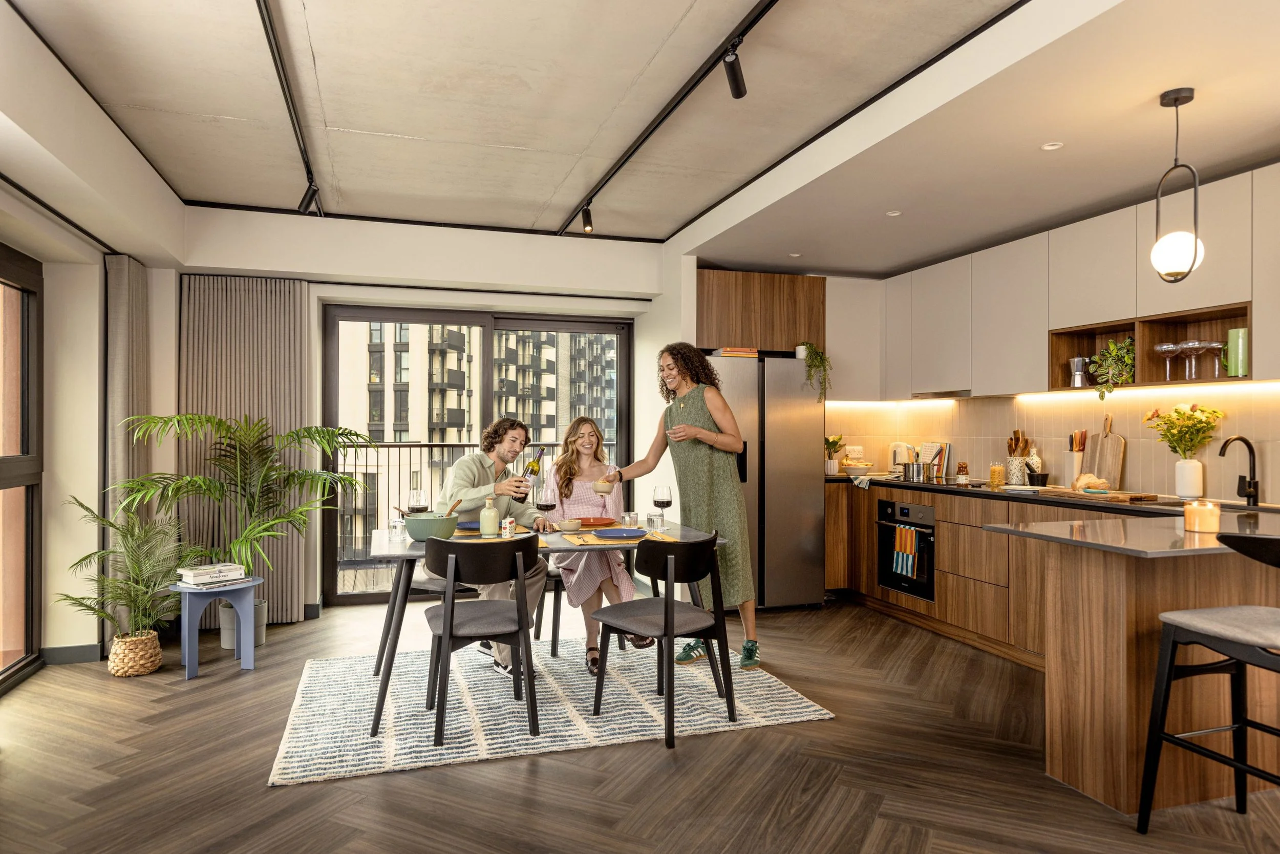 A modern kitchen and dining area with four people gathering around a dining table, enjoying a meal and drinks, with large windows, indoor plants, and contemporary decor.