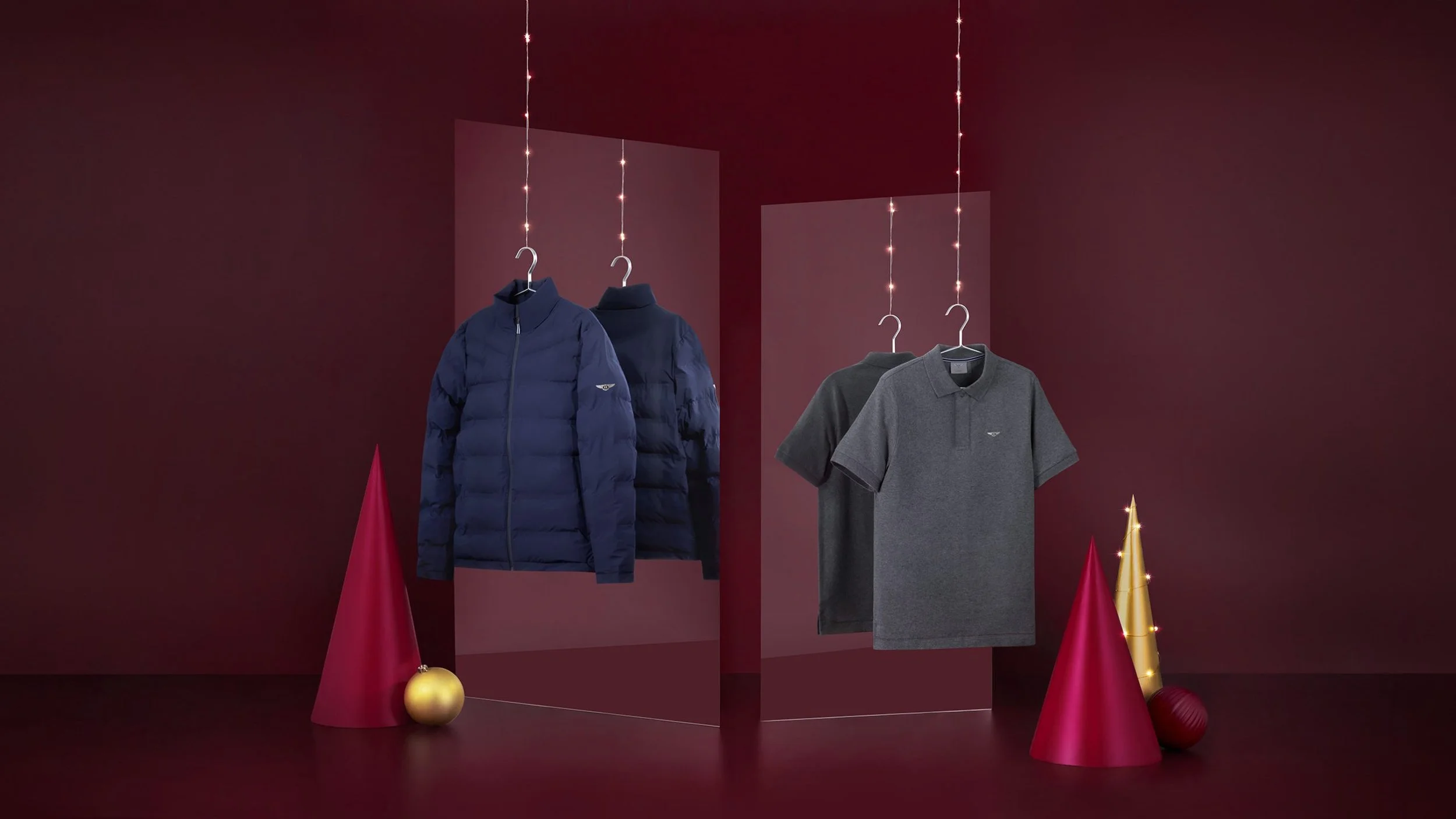 Display of men's clothing including a navy blue puffer jacket and a gray polo shirt, hanging on hangers against a dark red background with Christmas tree cone decorations and ornaments.