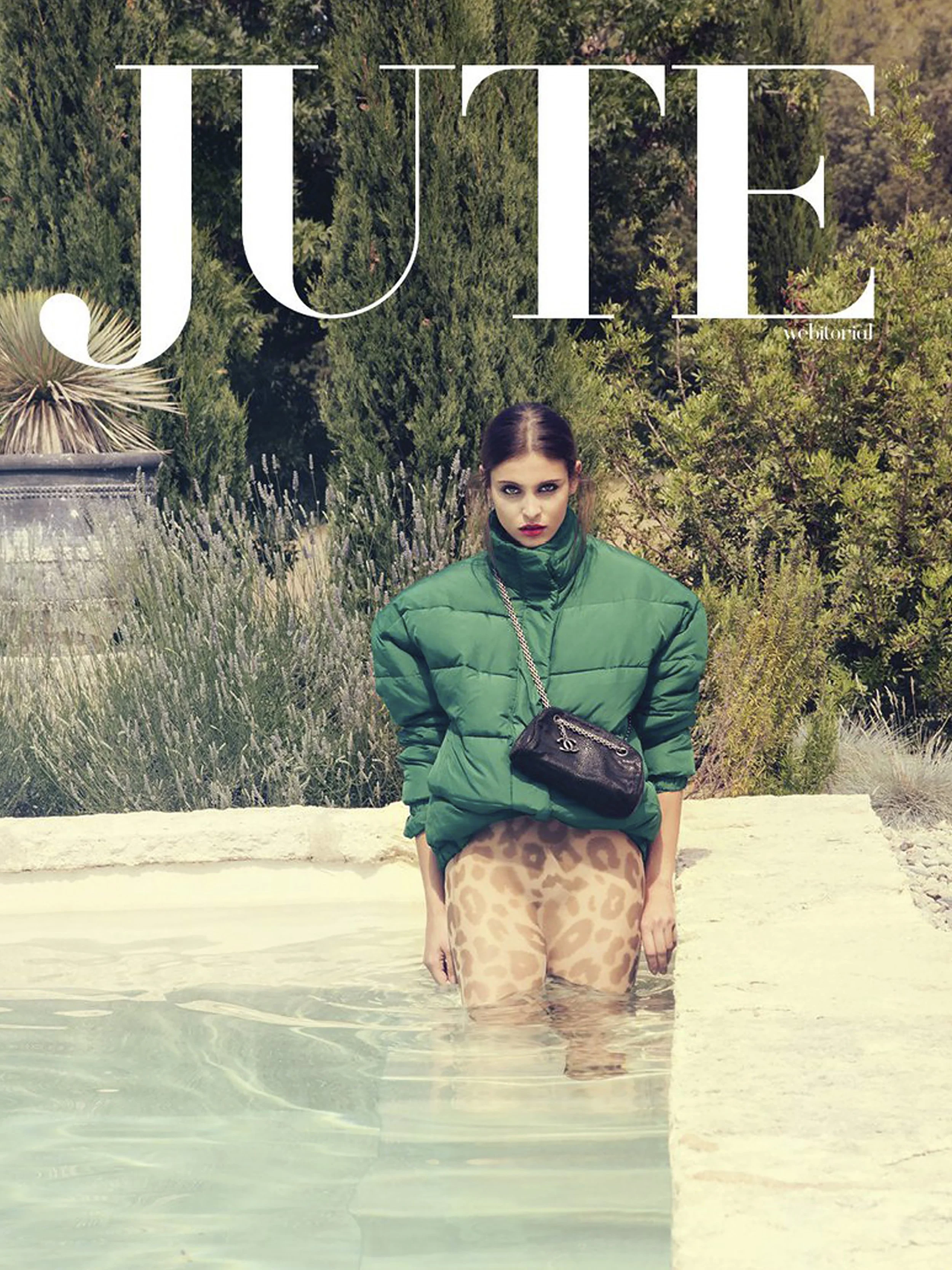 A woman with dark hair in a green puffer jacket and leopard print shorts standing in shallow water, with greenery behind her.