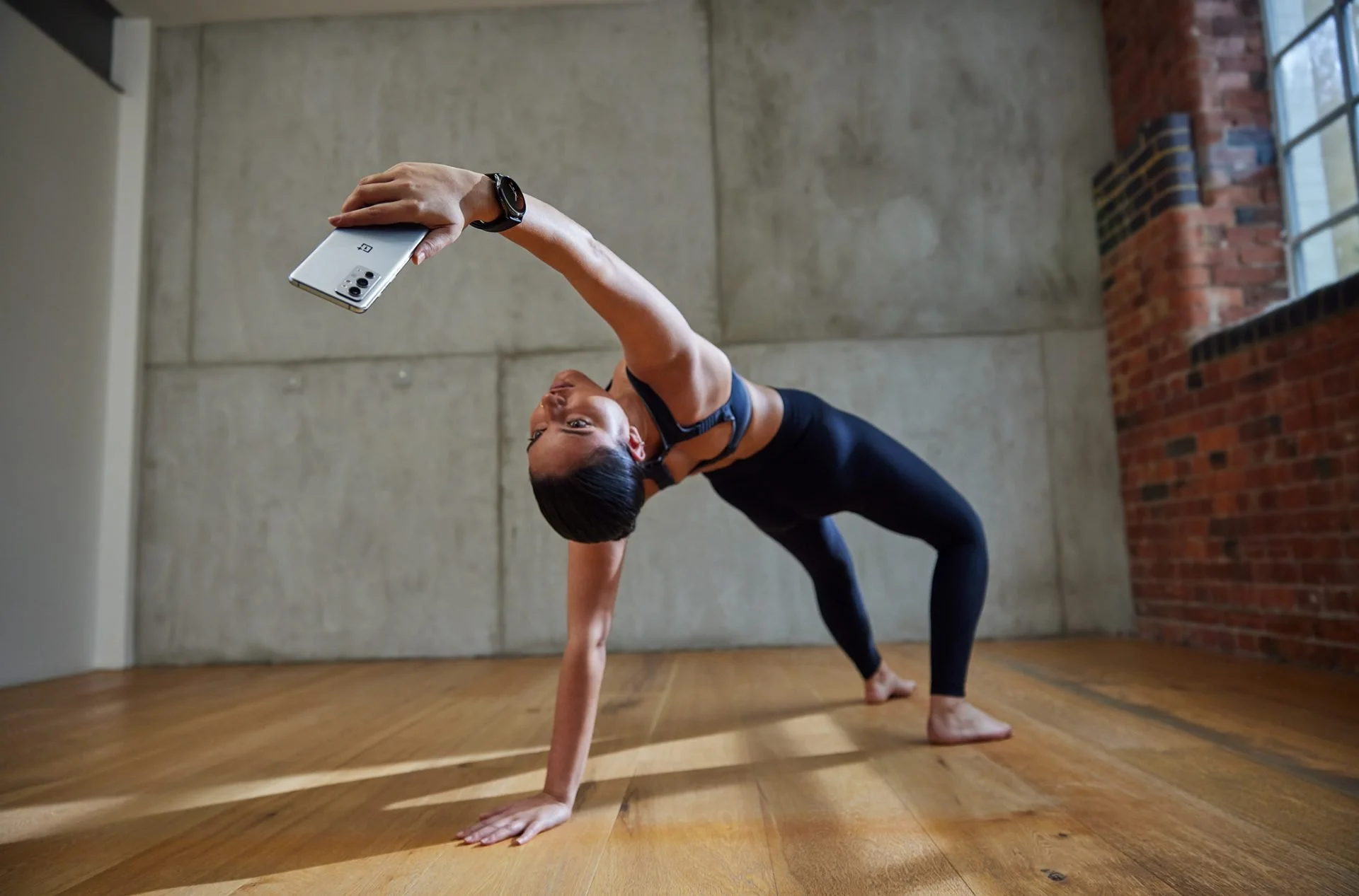 A woman performing a handstand yoga pose in a spacious room with a wooden floor, concrete walls, and a brick wall with window in the background.