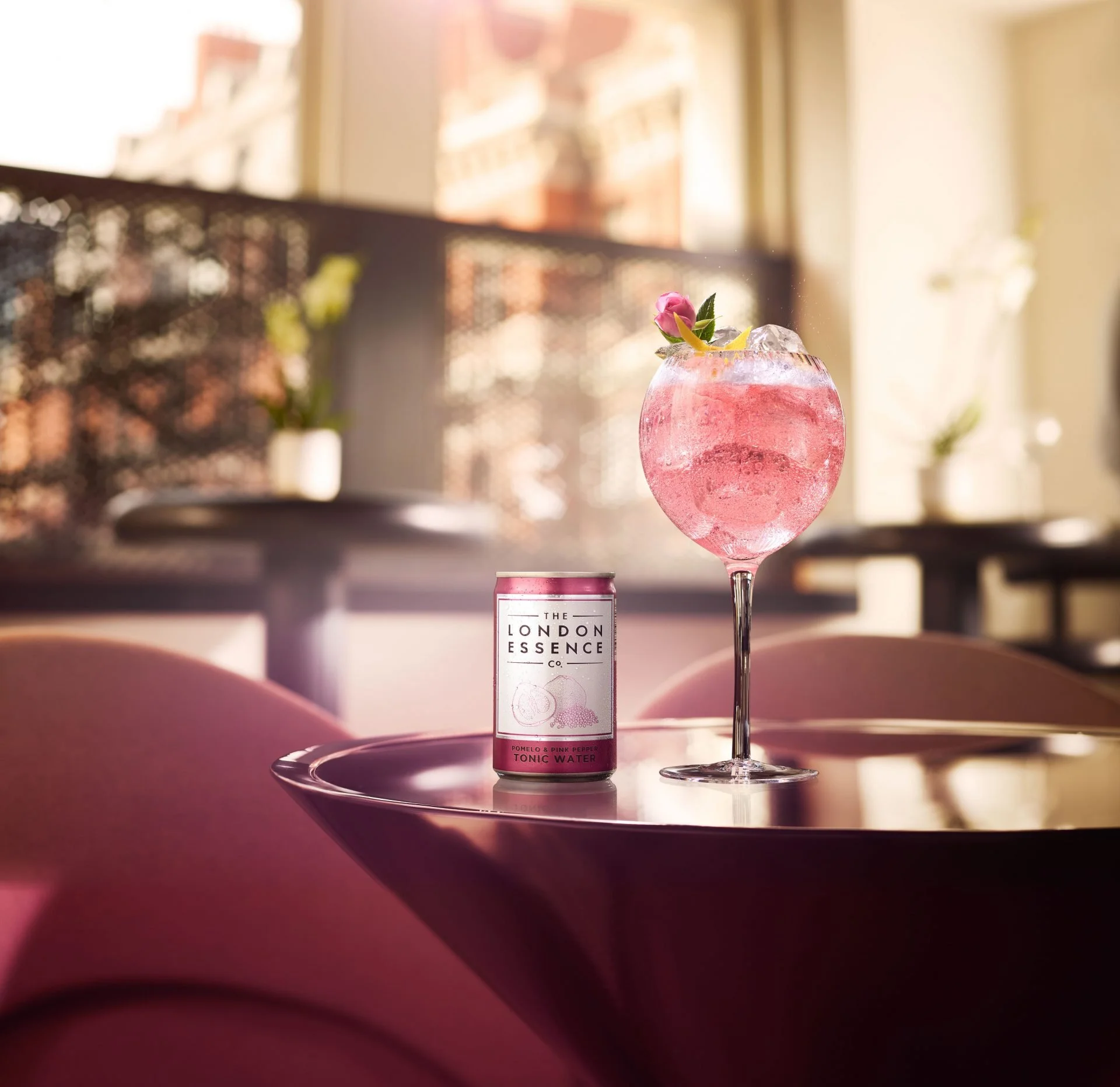 A pink cocktail with a small pink flower and lemon peel garnish in a rounded glass, placed on a pink tray. A bottle of sparkling tonic water labeled "The London Essence Co. Pomelo & Pink Pepper Tonic Water" is behind the glass. The scene is illuminat