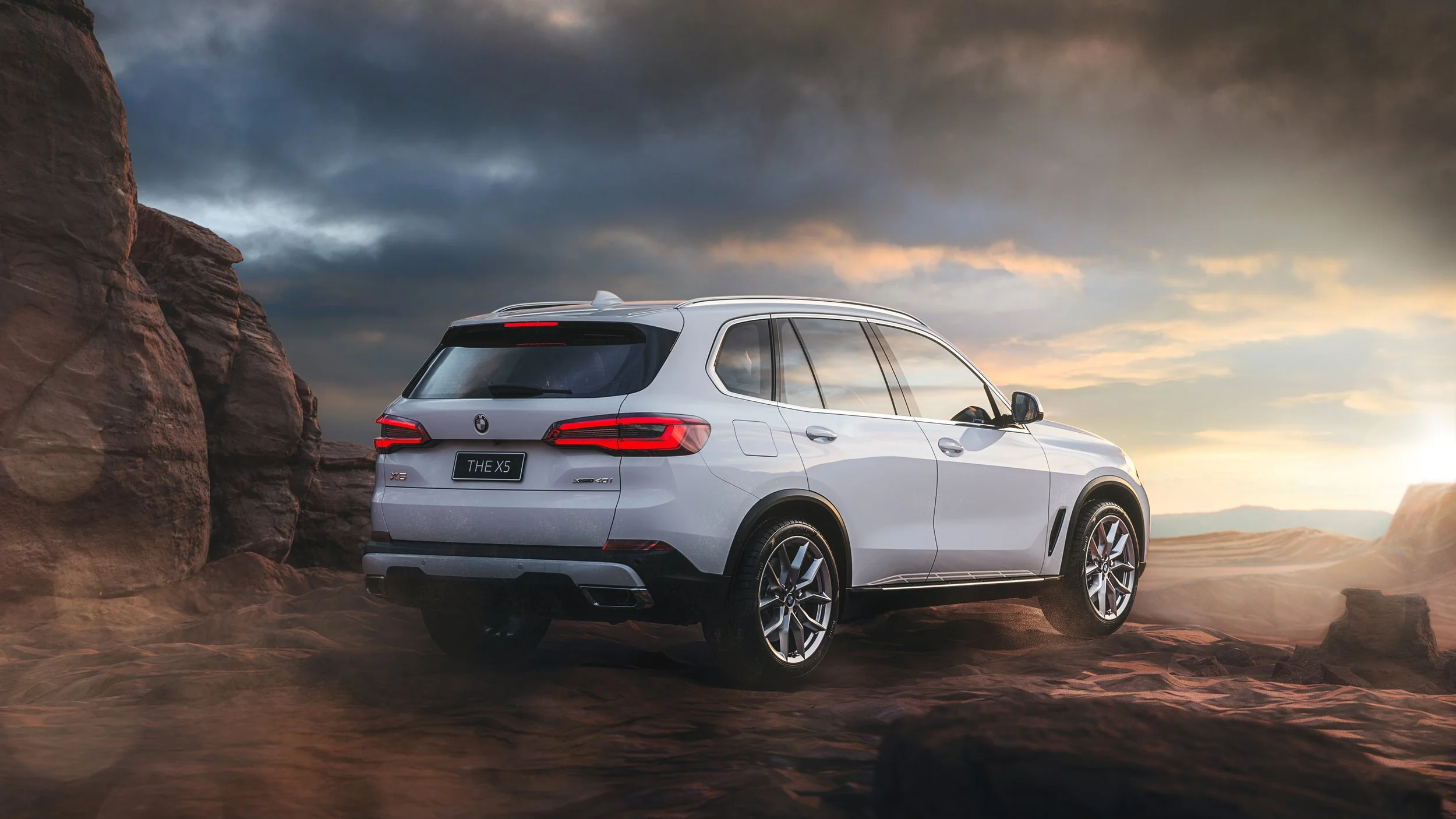 A white BMW X5 SUV parked on a rocky terrain with a dramatic sky of dark clouds and sunset in the background, near large desert rock formations.