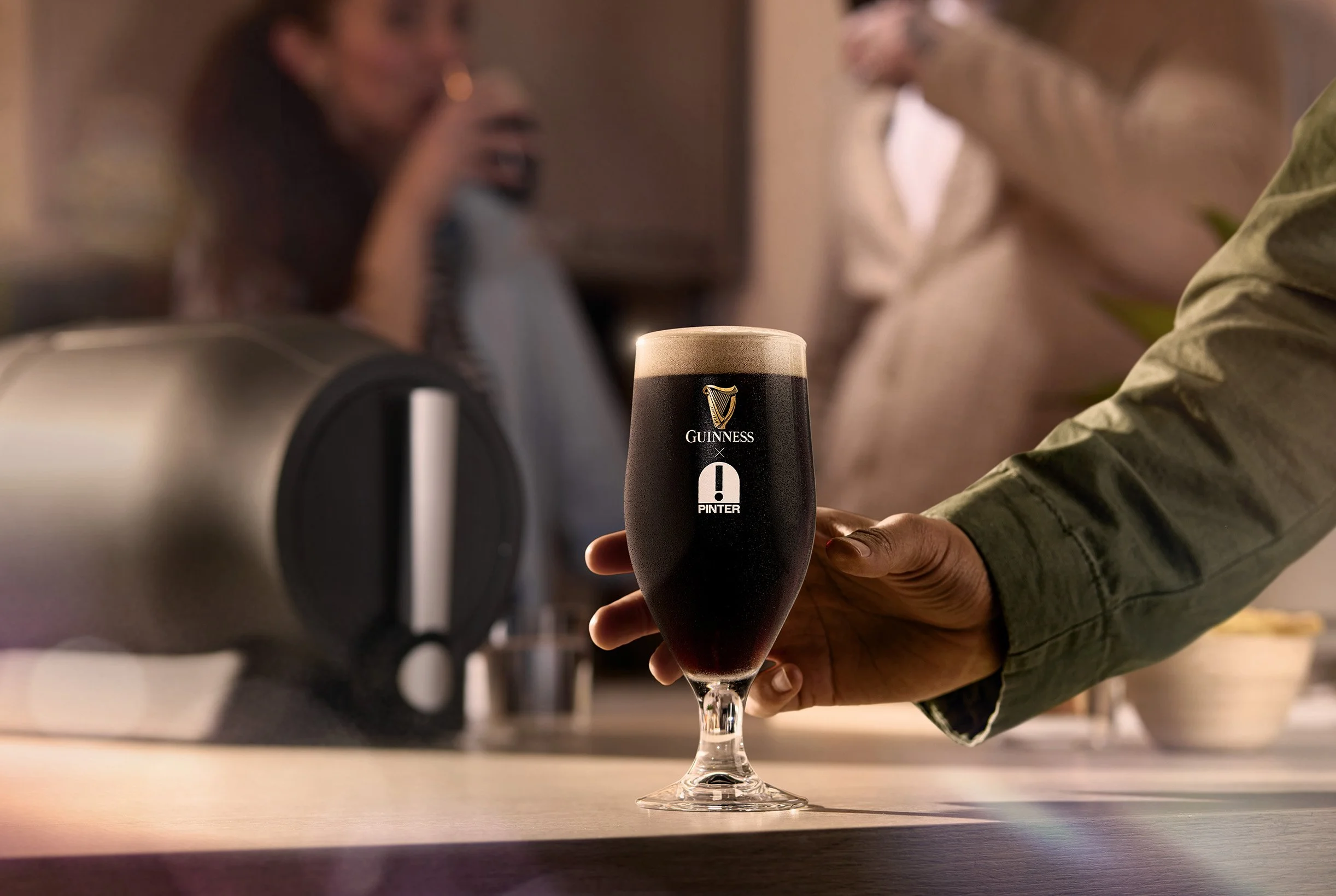 Person holding a pint glass of Guinness beer, with a foamy head, in a bar or pub setting.