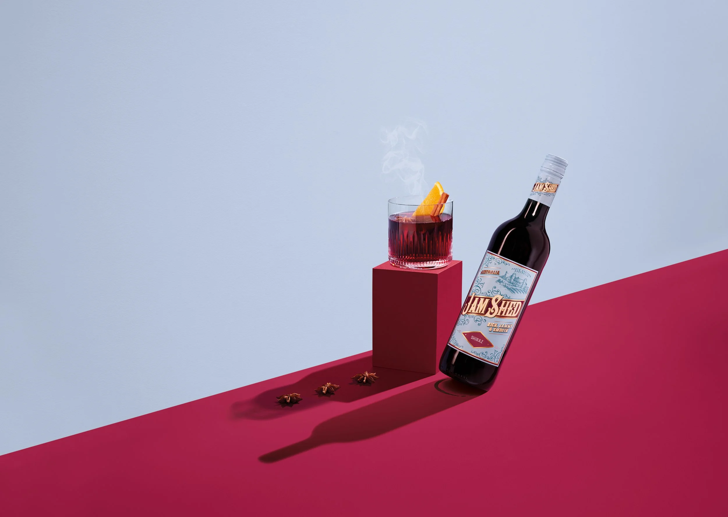 A glass of red wine with steam rising, garnished with an orange wedge and cinnamon stick, positioned on a red block. A bottle of Jam & Shred Shiraz wine leans against the block, with star anise spices and a red surface in the foreground against a pal