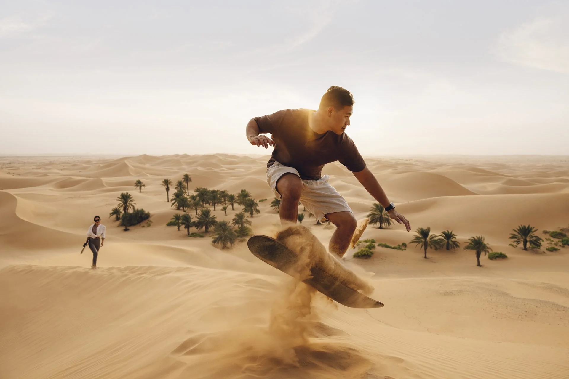 A man riding a skateboard on sand dunes in a desert landscape with palm trees, another person walking in the background, and a cloudy sky above.