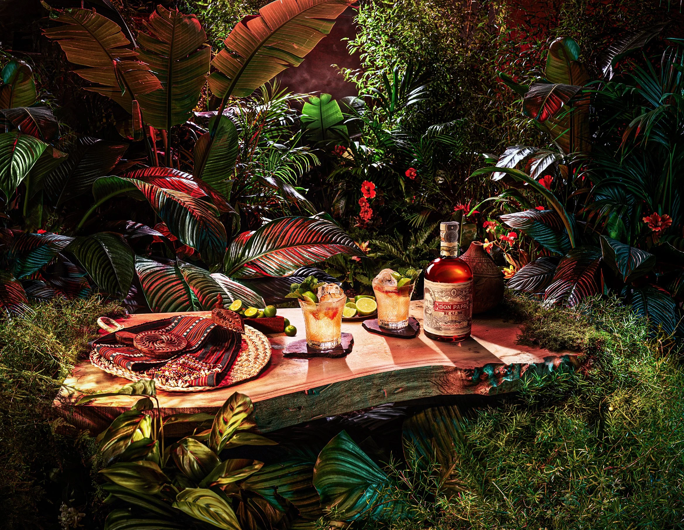 A tropical outdoor scene with lush green and red leaves, a table set with drinks, limes, a bottle of rum, and decorative items, creating a festive atmosphere.