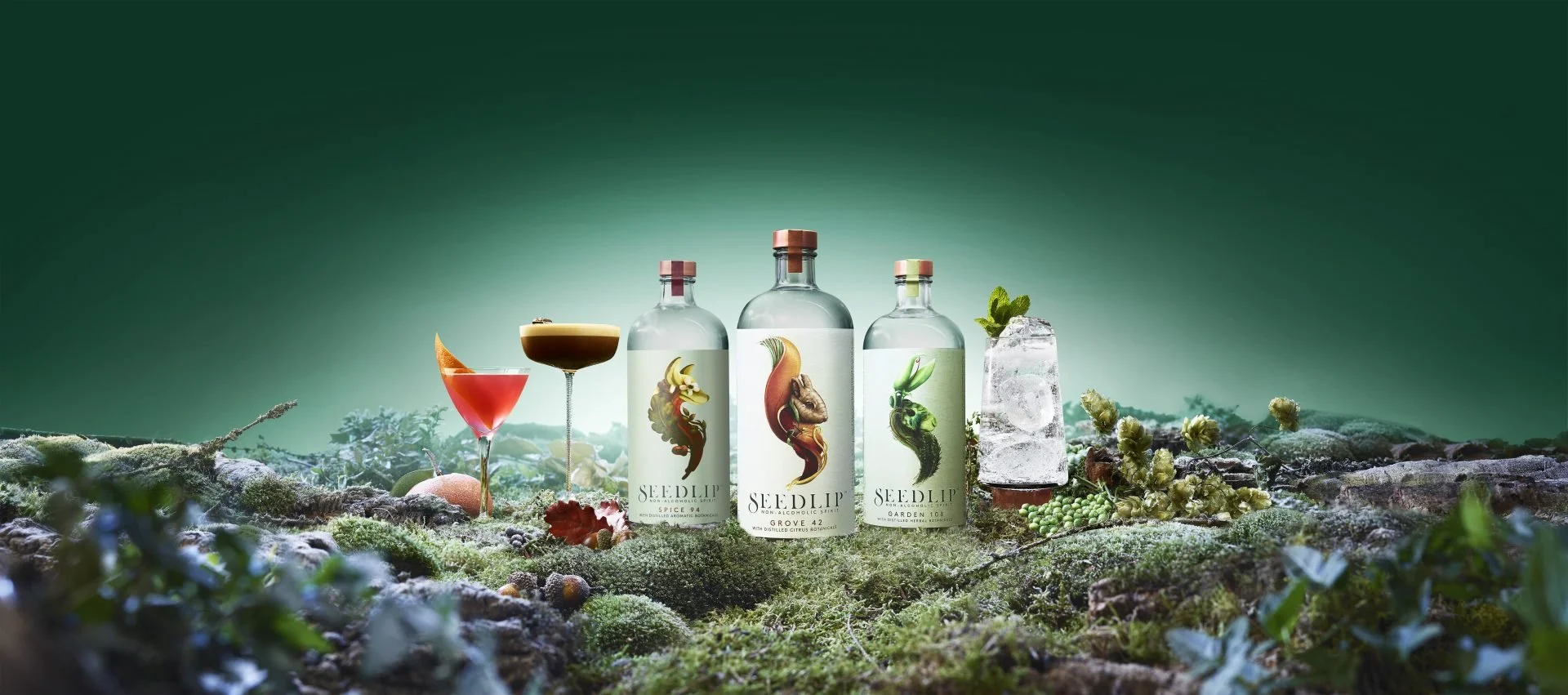 Display of non-alcoholic spirits and cocktails on a mossy surface with a green background, including bottles labeled Seedlip with botanical illustrations, a cocktail glass with a red drink and orange slice, a coupe glass with a dark drink, and a tall