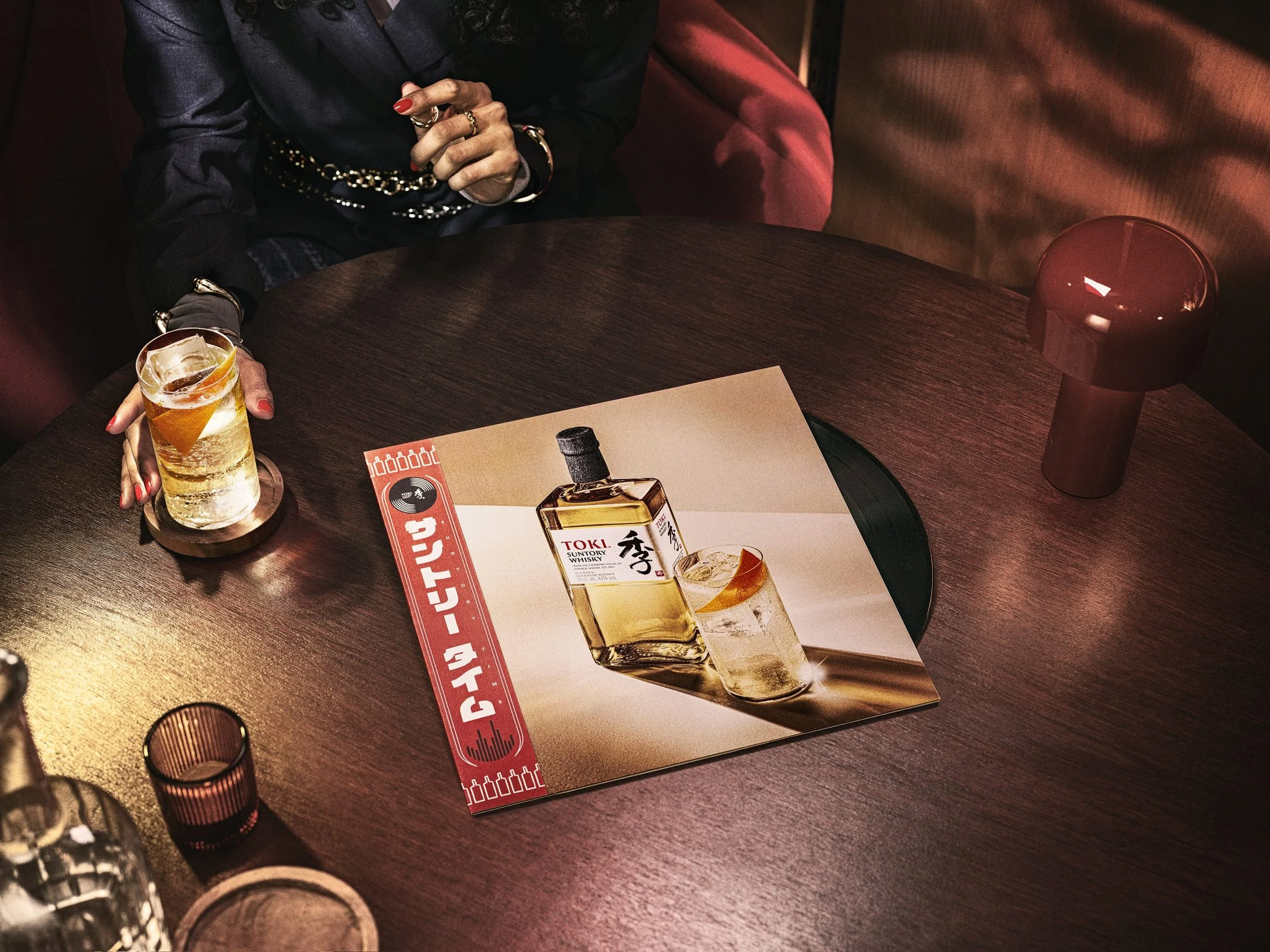 A person sitting at a wooden table with a glass of a golden beverage with ice and an orange slice, and a magazine featuring whiskey. The person's hands have rings, and they are wearing a black jacket with gold jewelry. There is a red lamp on the tabl