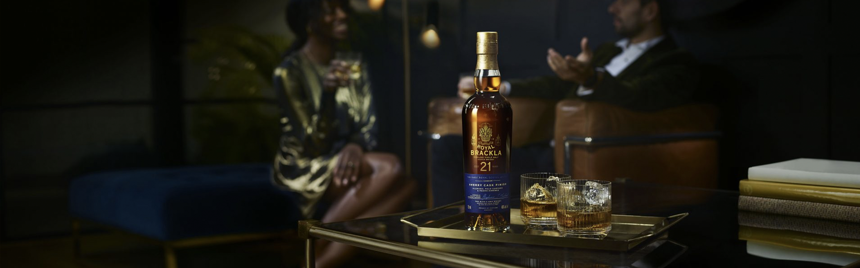 A bottle of Royal Brackla 21-year-old single malt Scotch whisky on a tray with two glasses filled with whisky and ice, set in a luxurious lounge with two people in the background.