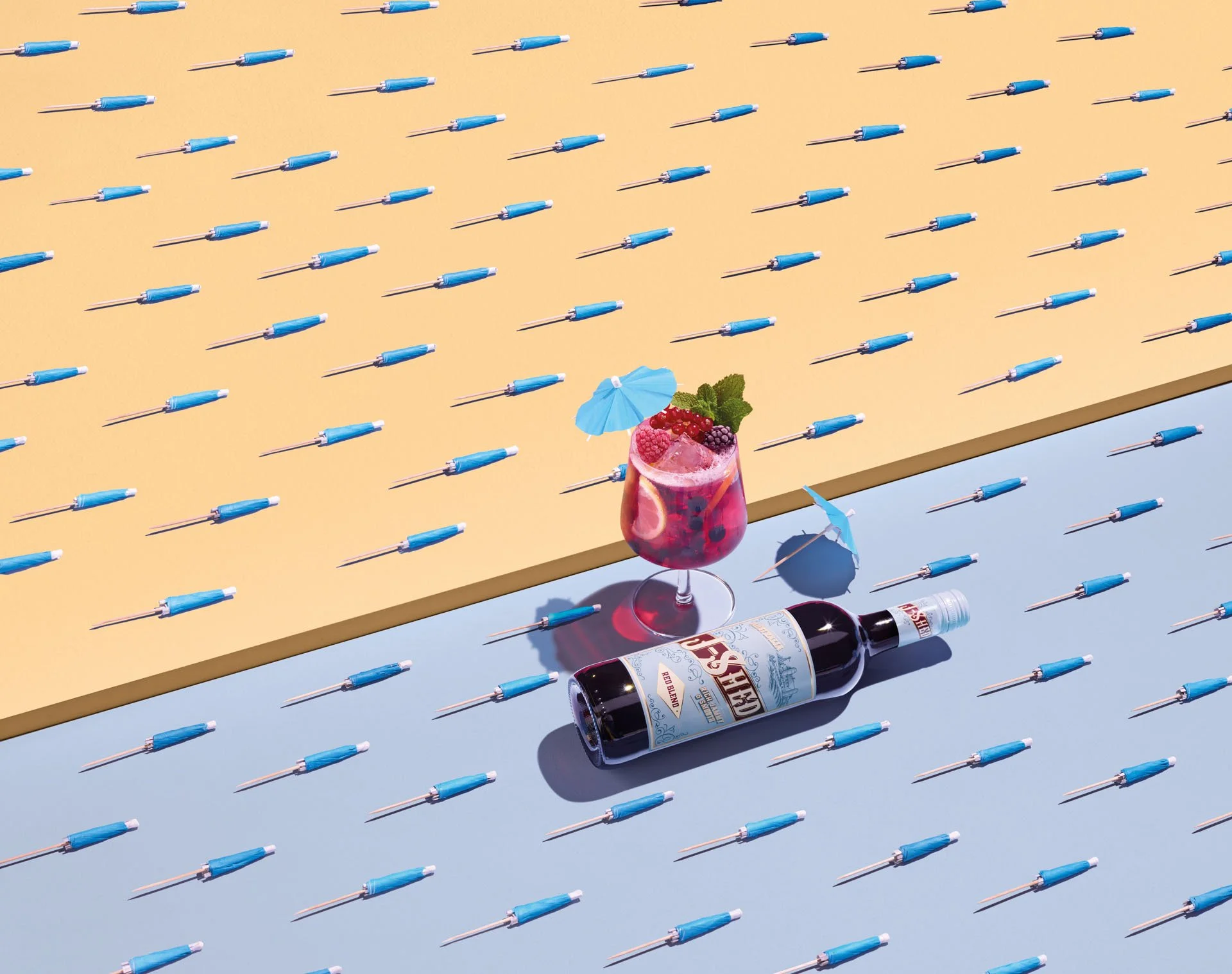 A yellow flat surface displays many small blue umbrellas and shadows. On the right side, core of a cocktail glass with pink drink, ice, berries, and a blue umbrella; nearby, a bottle of red wine labeled 'Red Bledzic' and a wine glass.