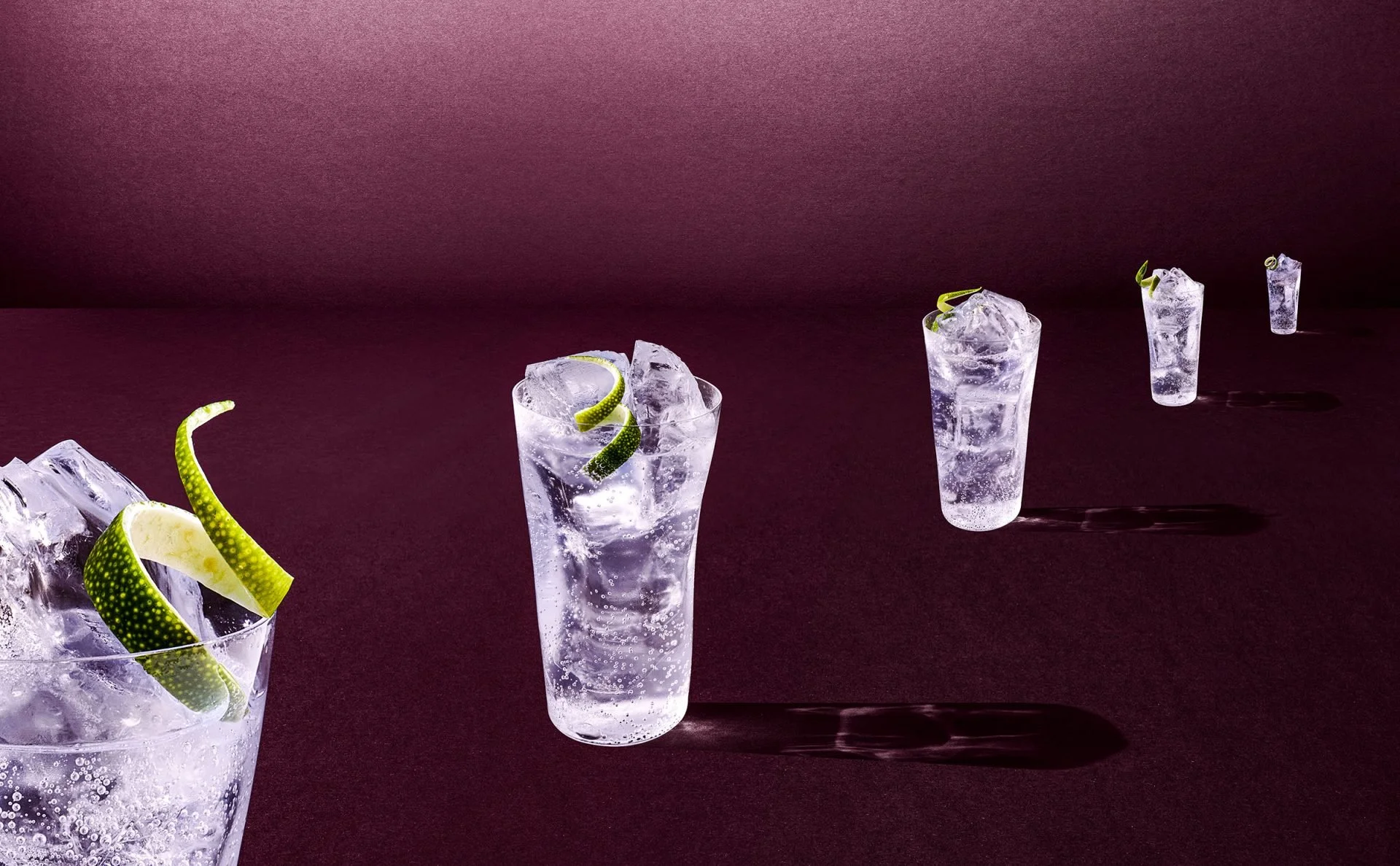 Multiple tall glasses filled with sparkling water and ice, garnished with lime twists, arranged on a dark surface against a dark pink background.