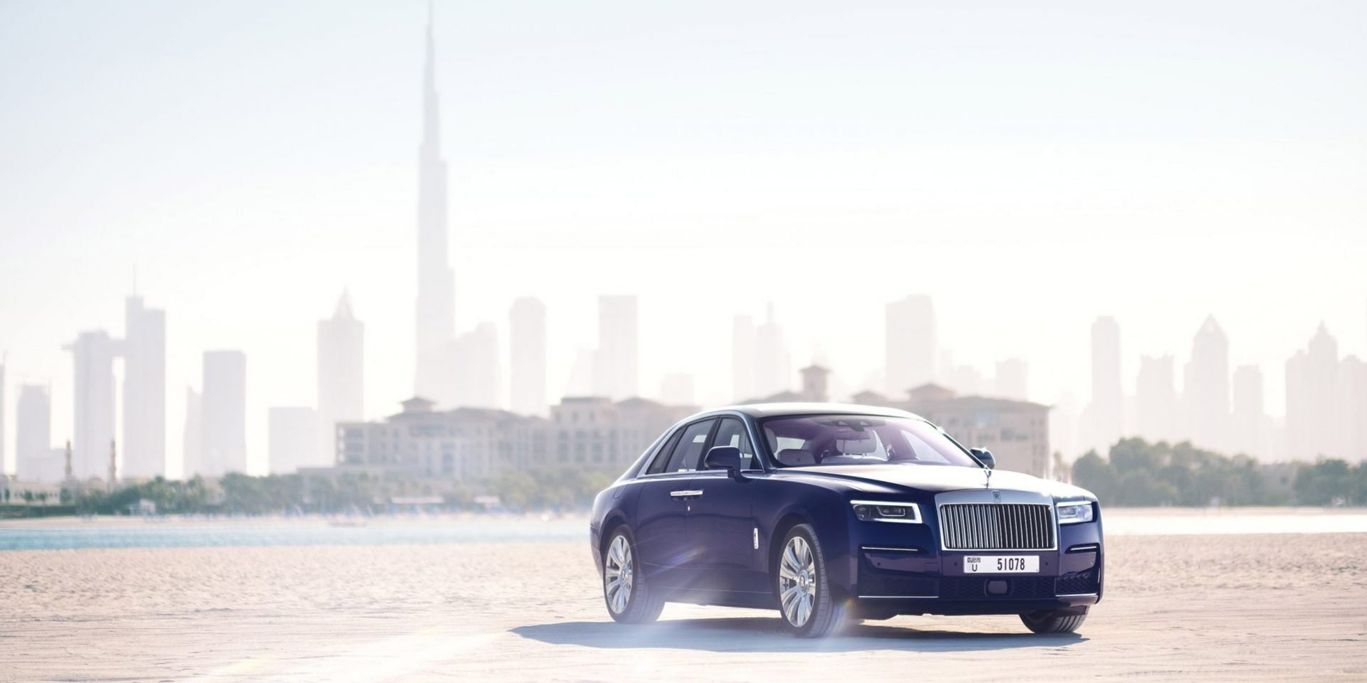 A luxurious black sedan car driving on a sandy beach with a city skyline in the background.