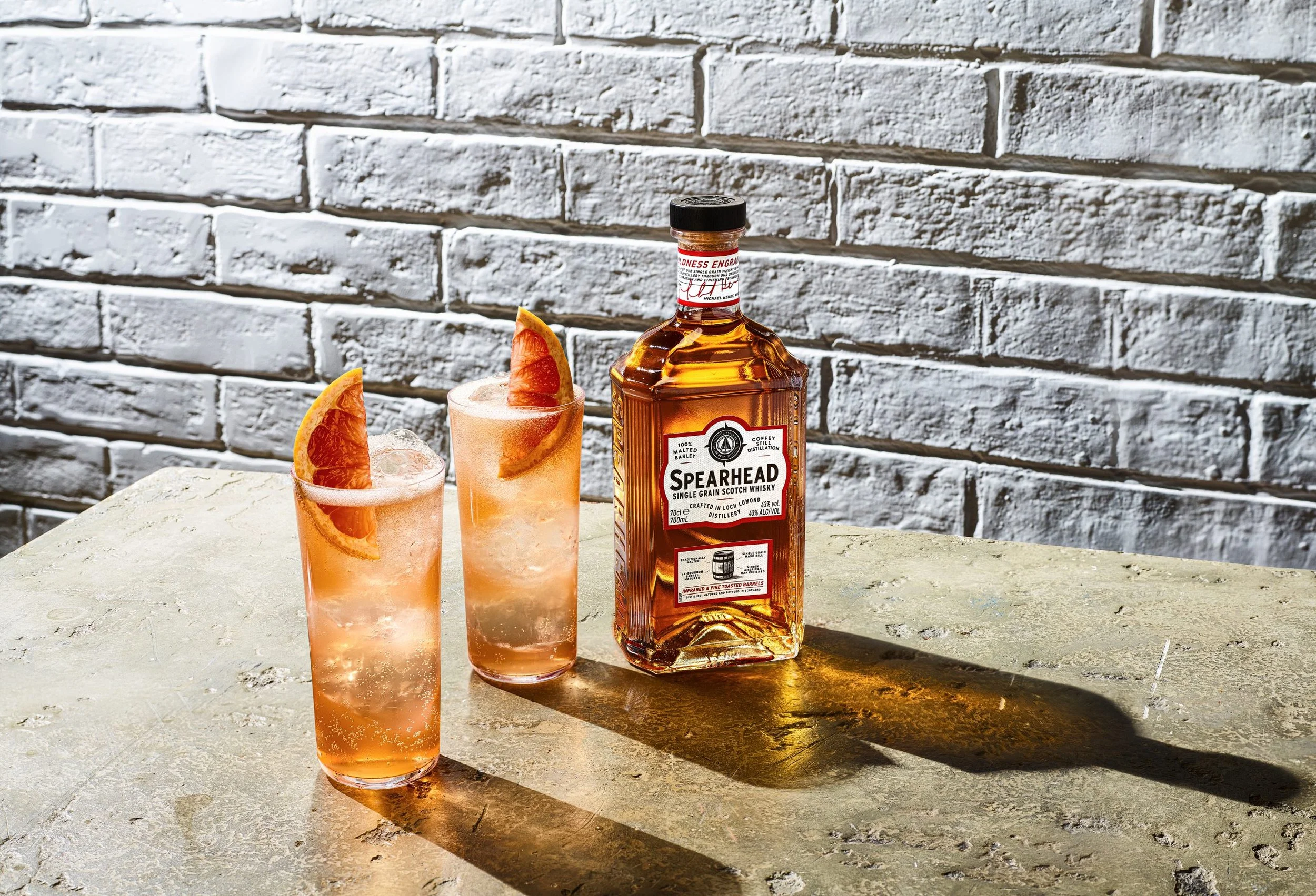 A bottle of Spearhead single grain scotch whisky and two pink cocktails garnished with orange slices on a concrete surface against a white brick wall