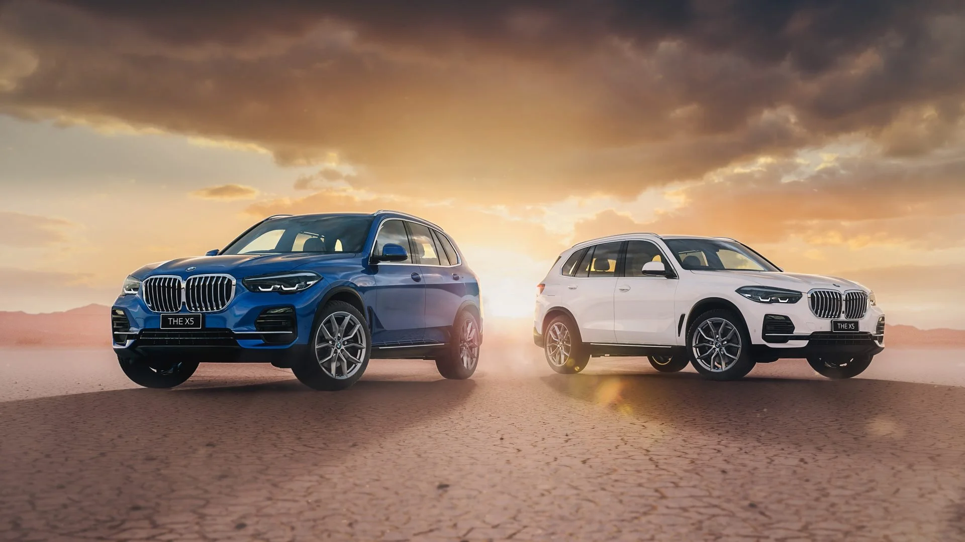 Two BMW X5 SUVs, one blue and one white, parked on desert terrain with a dramatic sunset in the background.