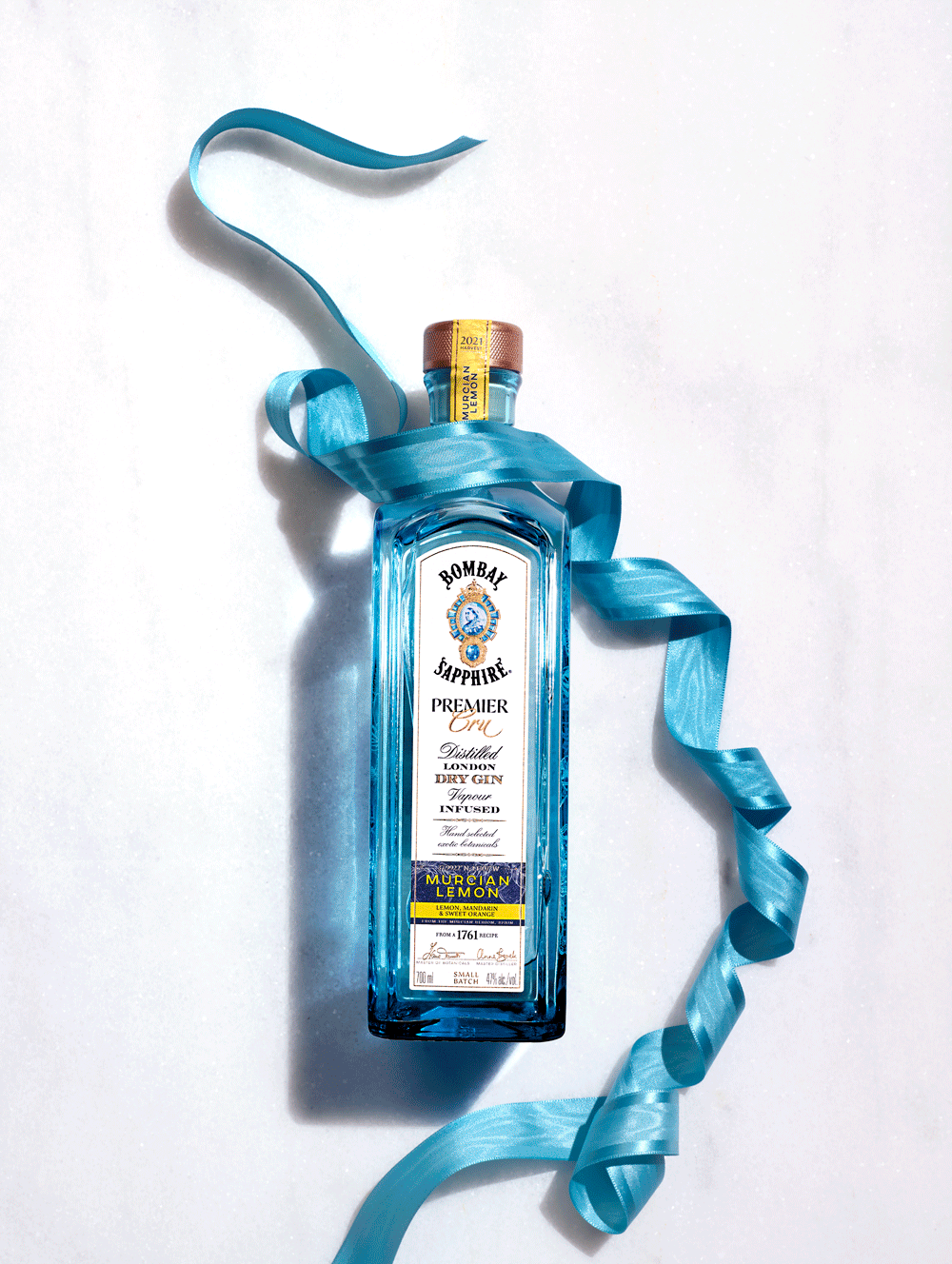 A blue-tinted bottle of Bombay Sapphire gin decorated with a blue ribbon, with a label indicating it is flavored with Murcian lemon, placed on a white background.