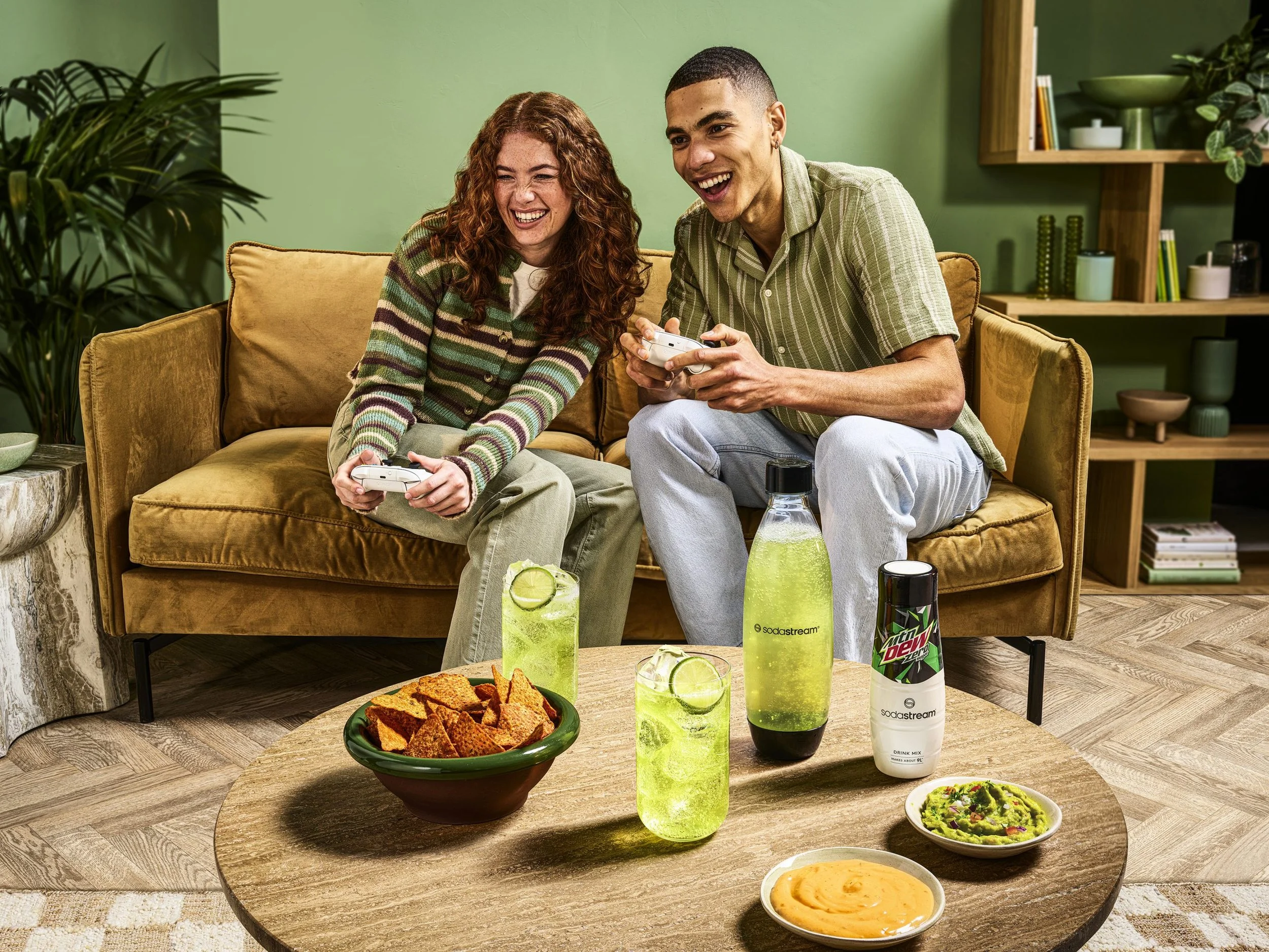 Two young adults, a woman with curly red hair and a man with short dark hair, sitting on a yellow couch in a living room, playing video games together and smiling.