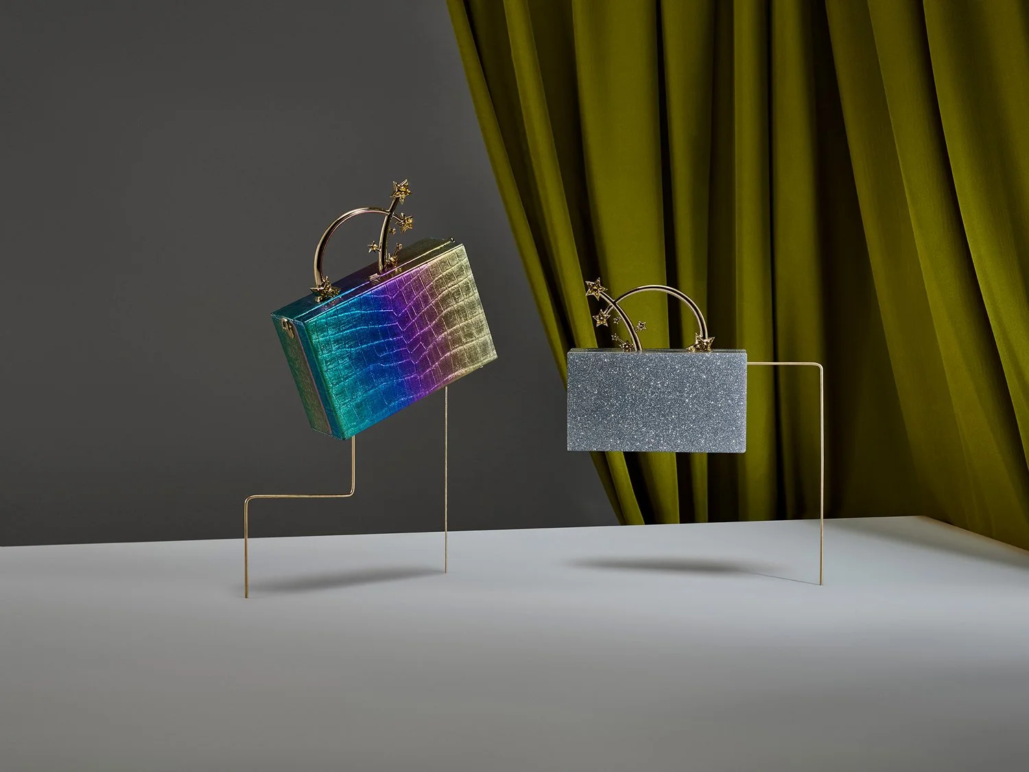 Two stylish handbags with star and arc details are displayed on minimalist gold stands in front of a dark background and green curtains.