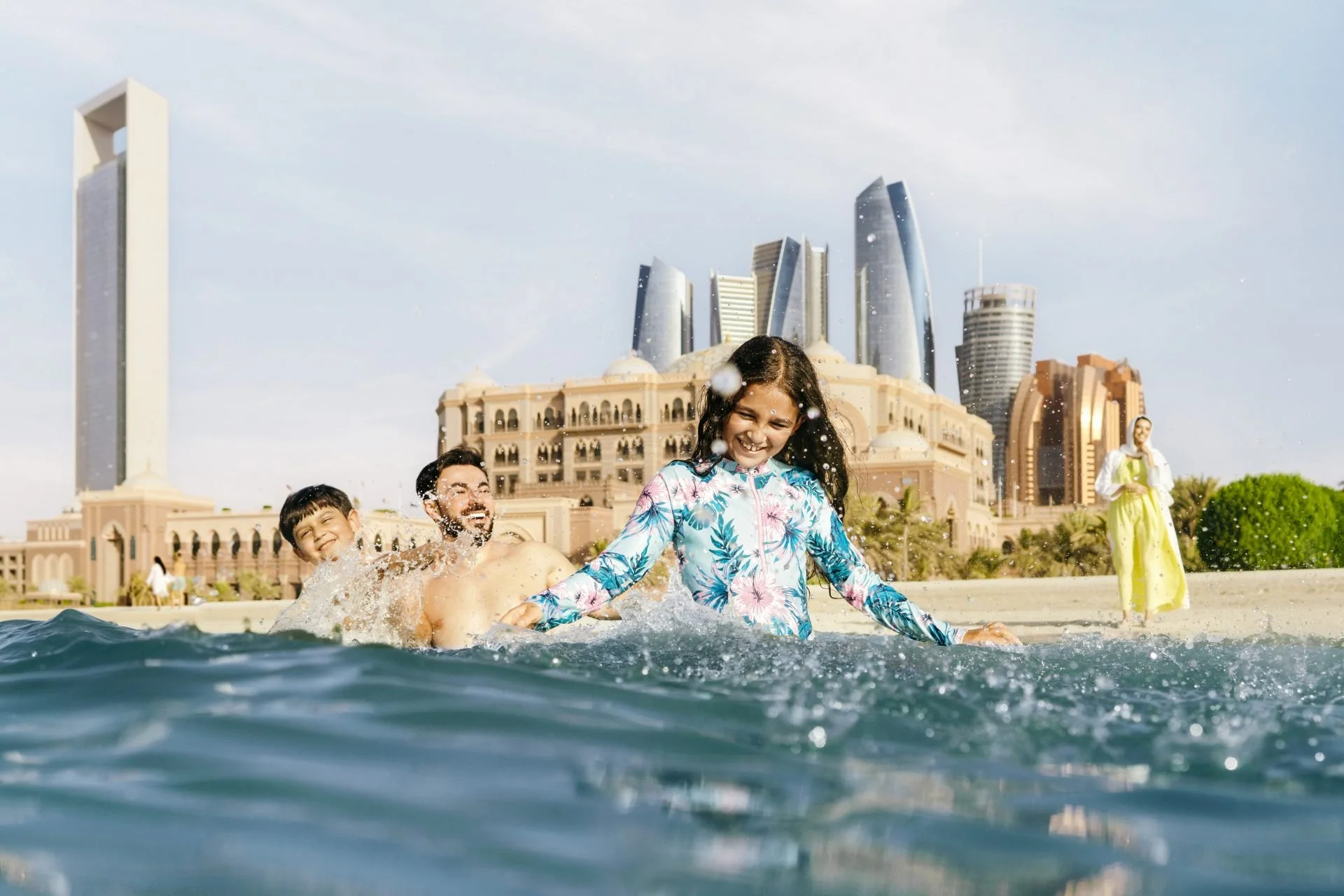 Children playing and splashing in a pool in front of a luxurious hotel with modern skyscrapers in the background.