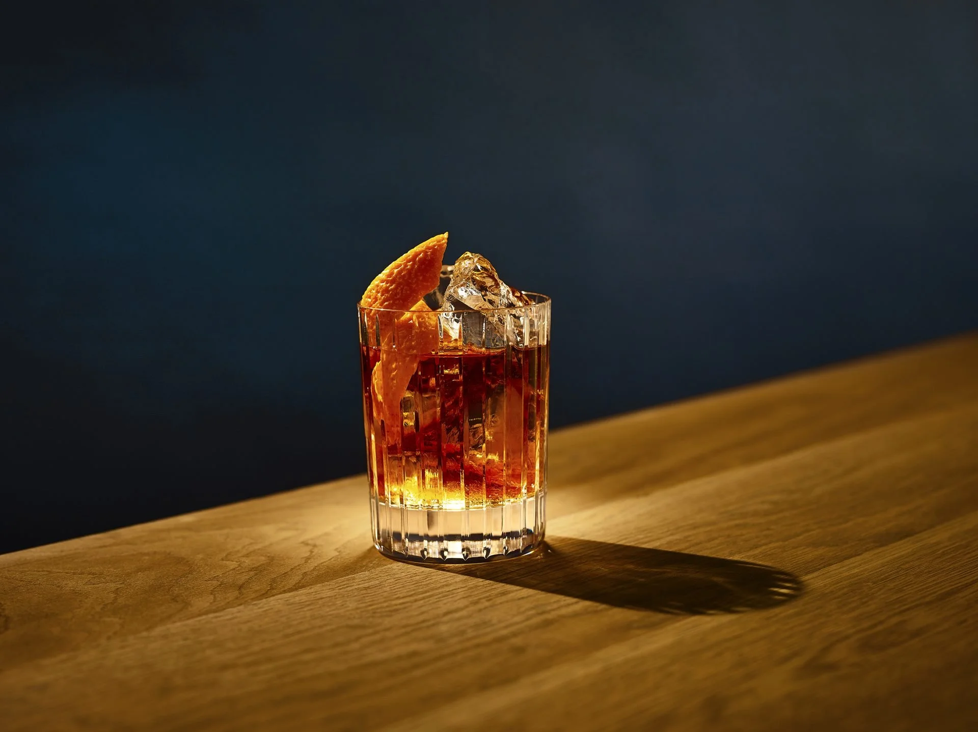 A glass of cocktail with ice and an orange wedge on a wooden surface, lit by warm light.