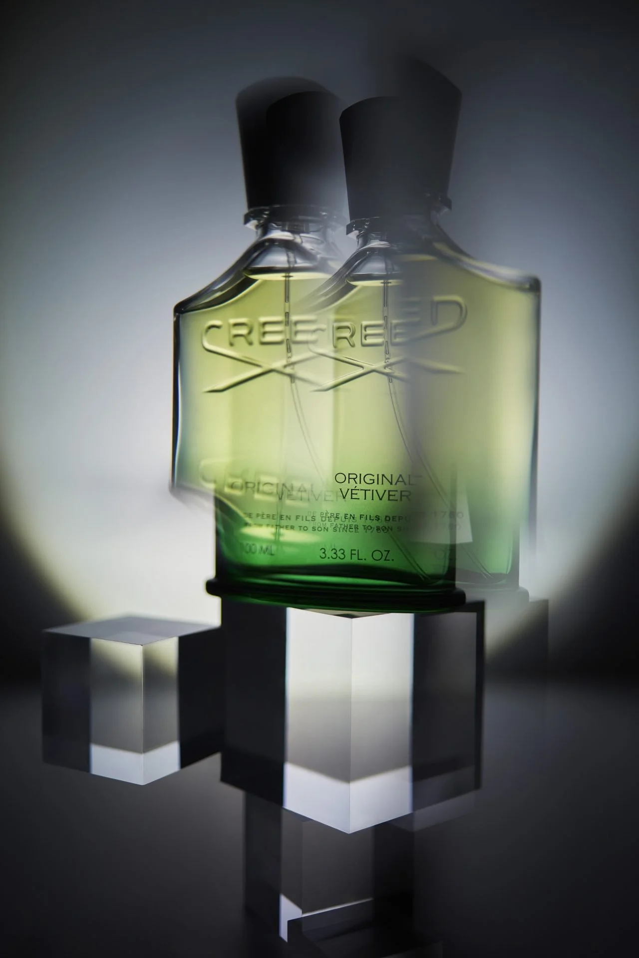 A perfume bottle with light green liquid, embossed with the brand name 'CREED' and the words 'ORIGINAL VÉTIVER,' displayed on a clear stand and surrounded by reflective cube-shaped objects.