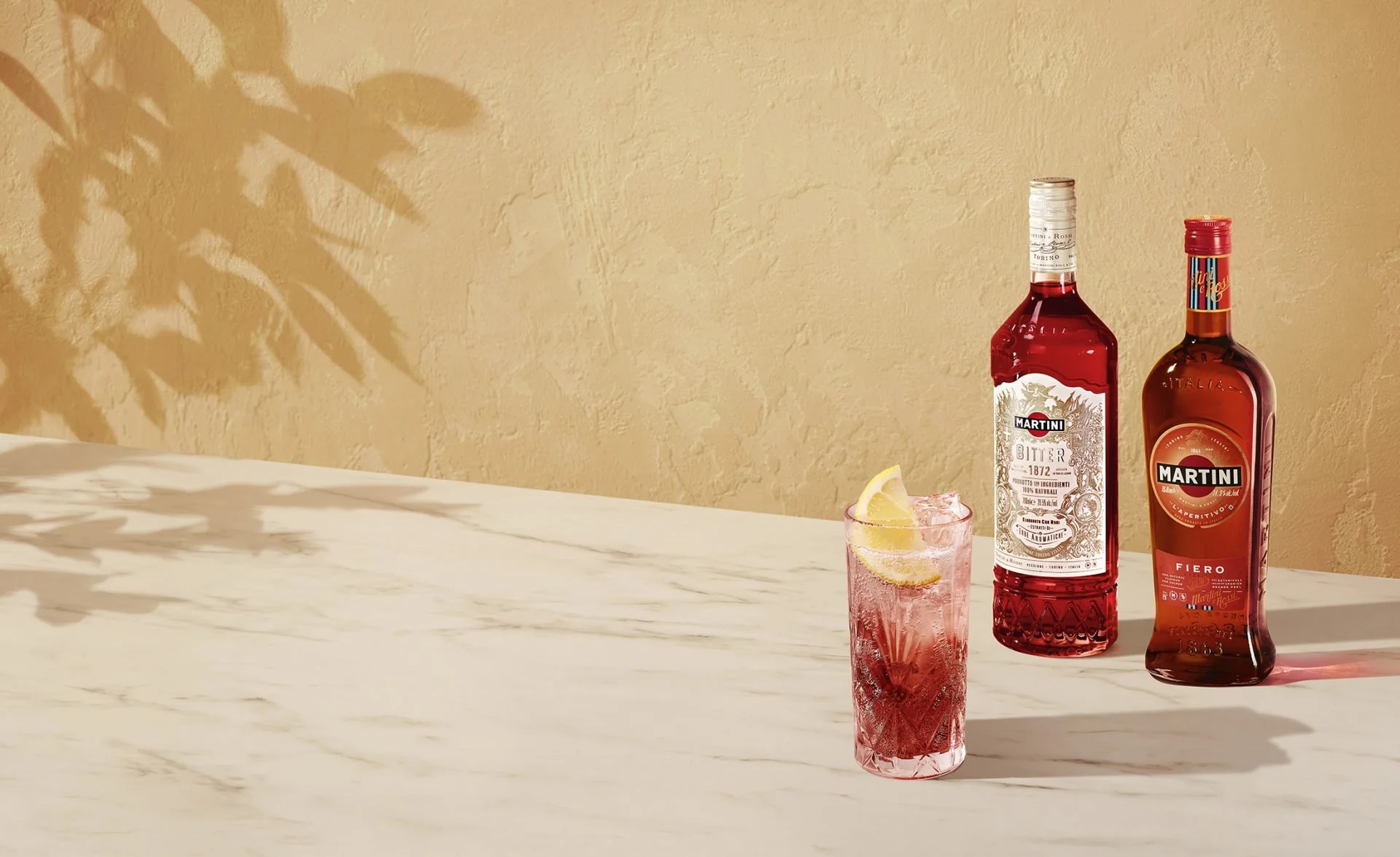 A marble countertop with two bottles of Martini liquor, a pink cocktail with lemon slices, and a beige textured wall with a shadow of leaves.