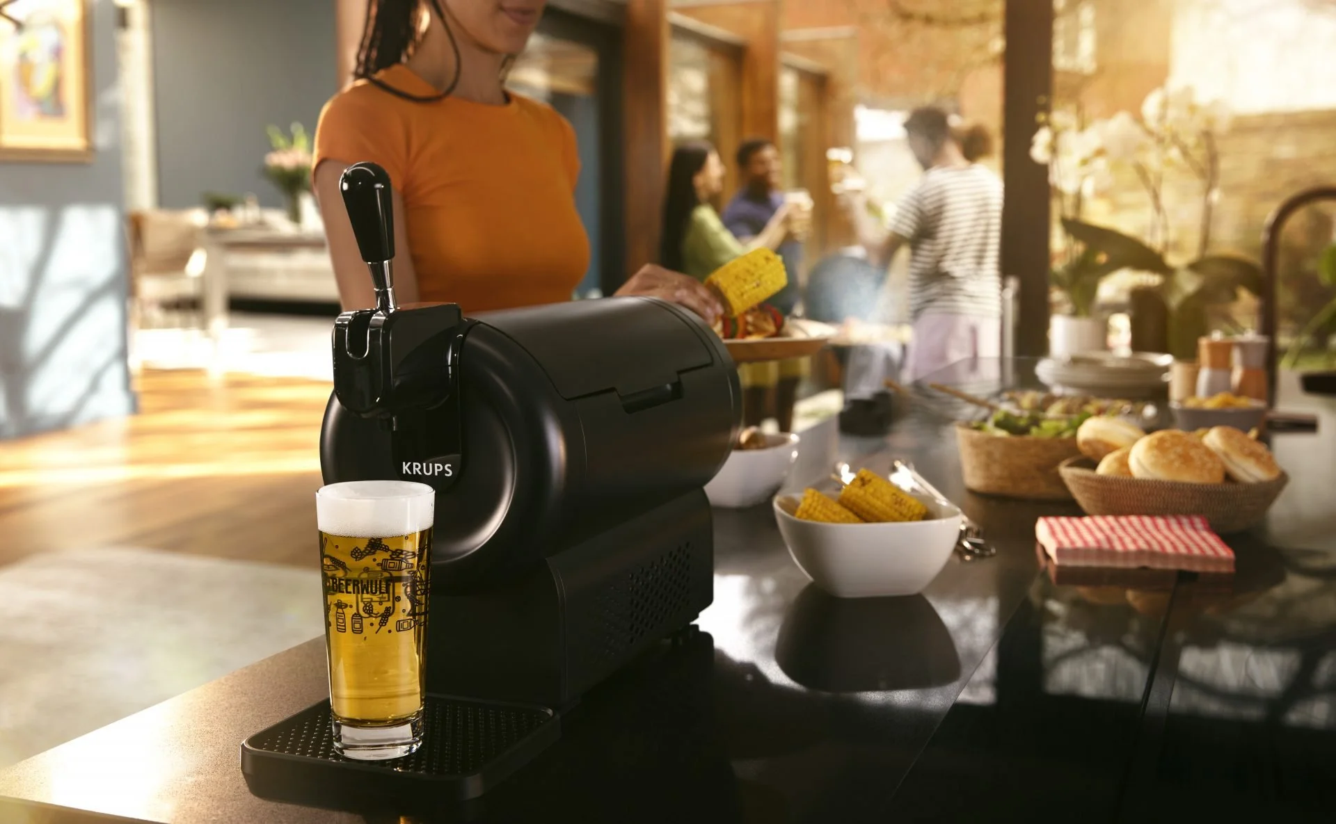 A woman in an orange shirt standing at a buffet table with a black Krups beverage dispenser and a glass of beer next to it. The table has bowls of corn on the cob, bread rolls, and various dishes. Other people are in the background in a well-lit room