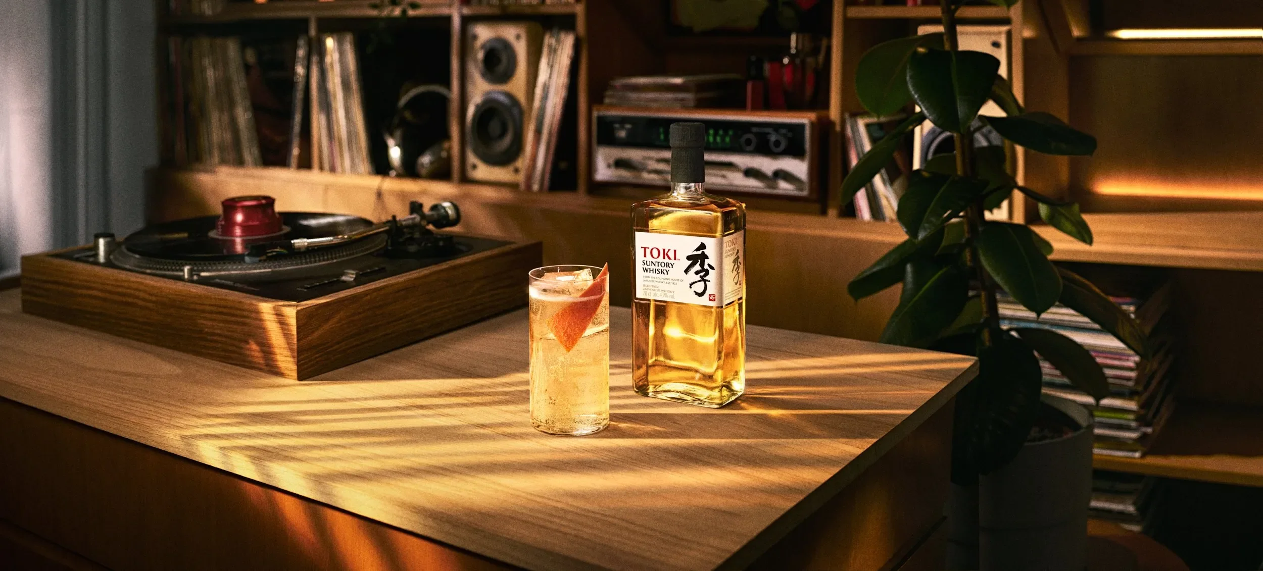 A wooden table with a bottle of Suntory Whisky, a glass with a drink and ice, a vinyl record player, and a potted plant in a cozy room with shelves of books and vinyl records.