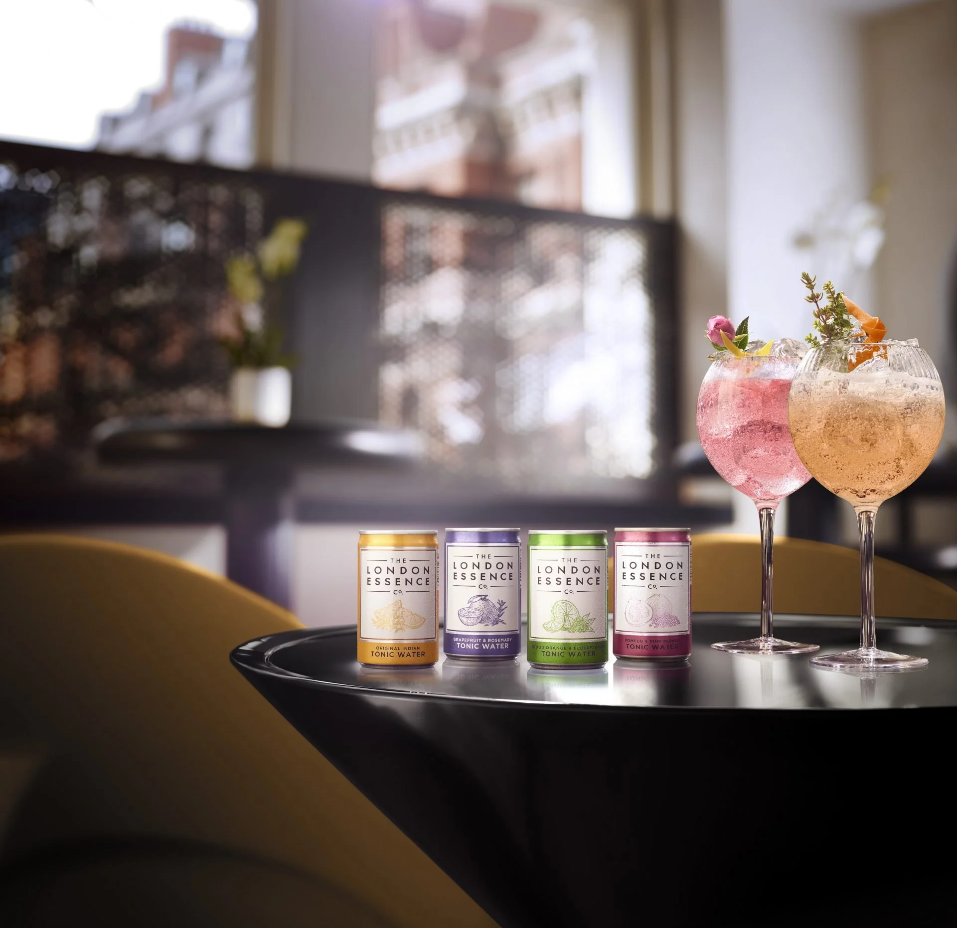 Four cans of tonic water with different flavors displayed on a black tray in front of a window with natural light, with pink and yellow cocktails garnished with flowers and citrus slices in the background.