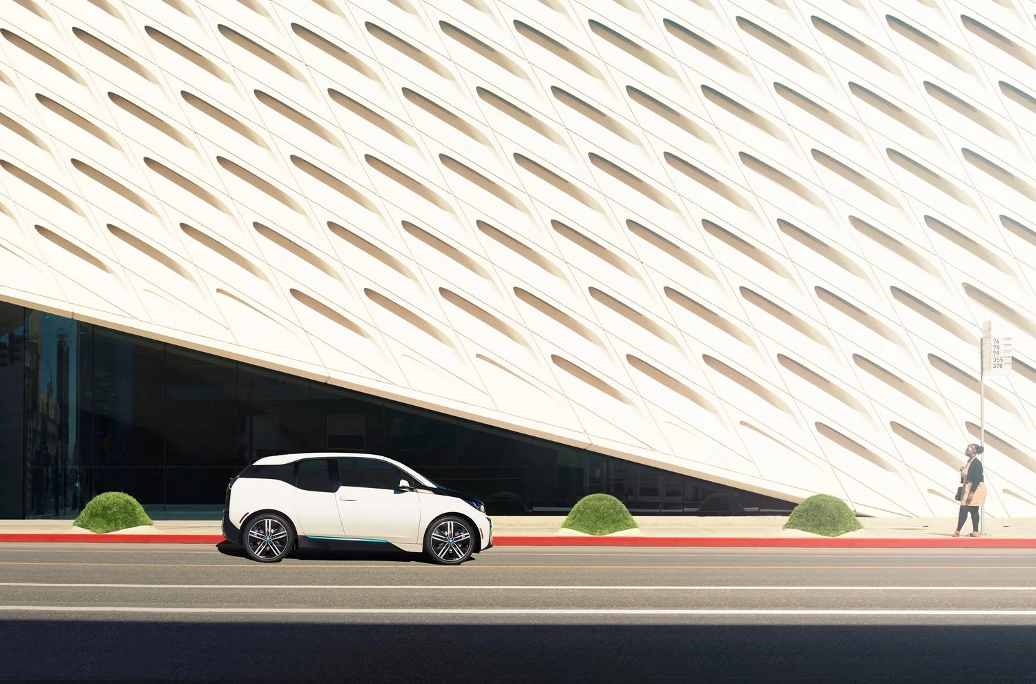 A white electric SUV parked in front of a modern building with a geometric facade. A person is walking on the sidewalk next to the building, and small green bushes are placed along the curb.