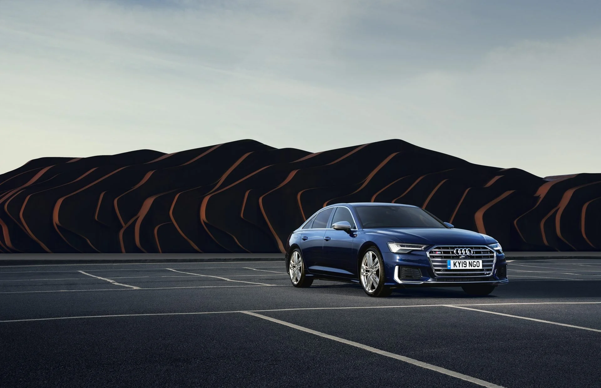 A dark blue Audi sedan parked in a nearly empty parking lot with white boundary lines, with a stylized mountain landscape in the background, under a cloudy sky.